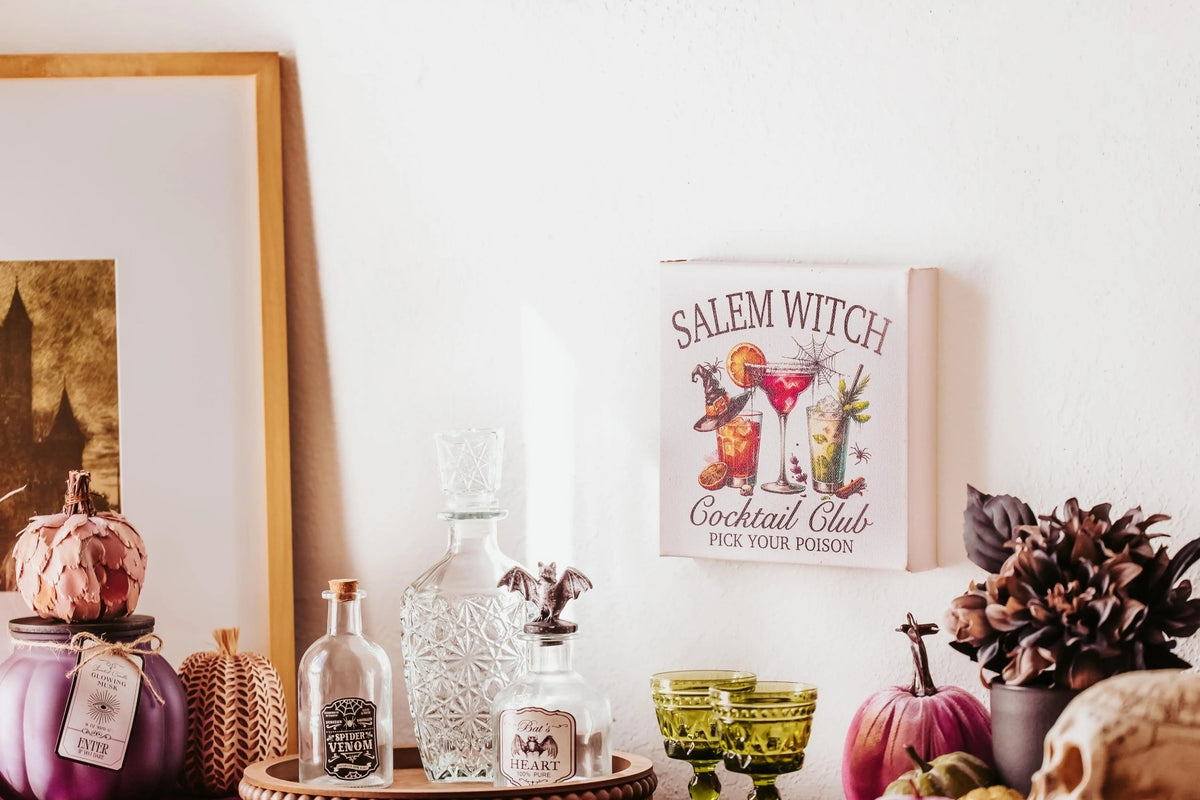 The Salem Witch Cocktail Club Halloween Canvas (8x8, Mirabilia Boutique) pops against a white wall—perfect spooky bar decor to create a witchy gallery wall vibe with vintage bottles, green glasses, and small pumpkins.