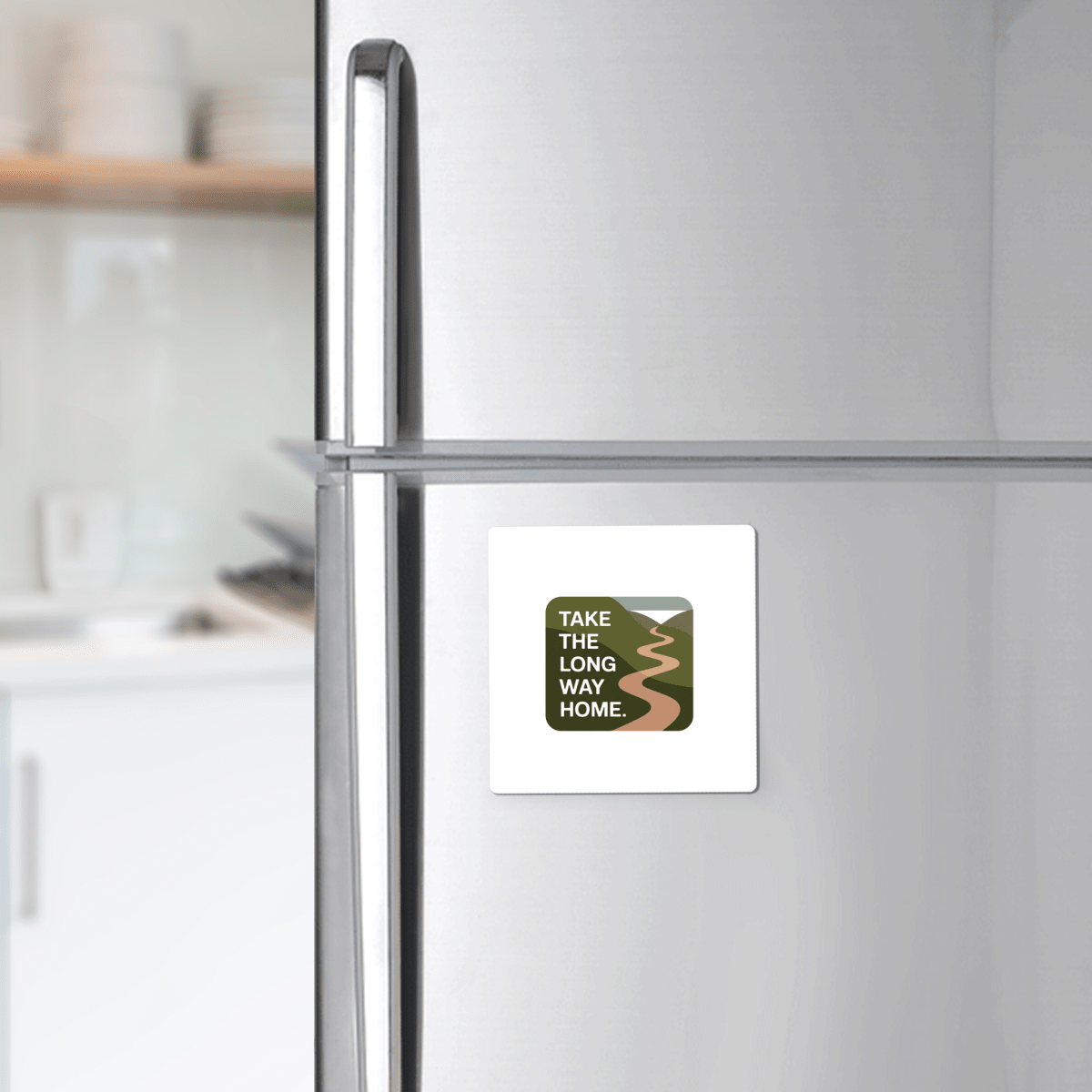 The Mirabilia Boutique "Take the Long Way Home Travel Magnet" is displayed on a stainless steel fridge, featuring a winding road illustration and text, set against nature-inspired décor with white dishes on shelves in a blurred kitchen.