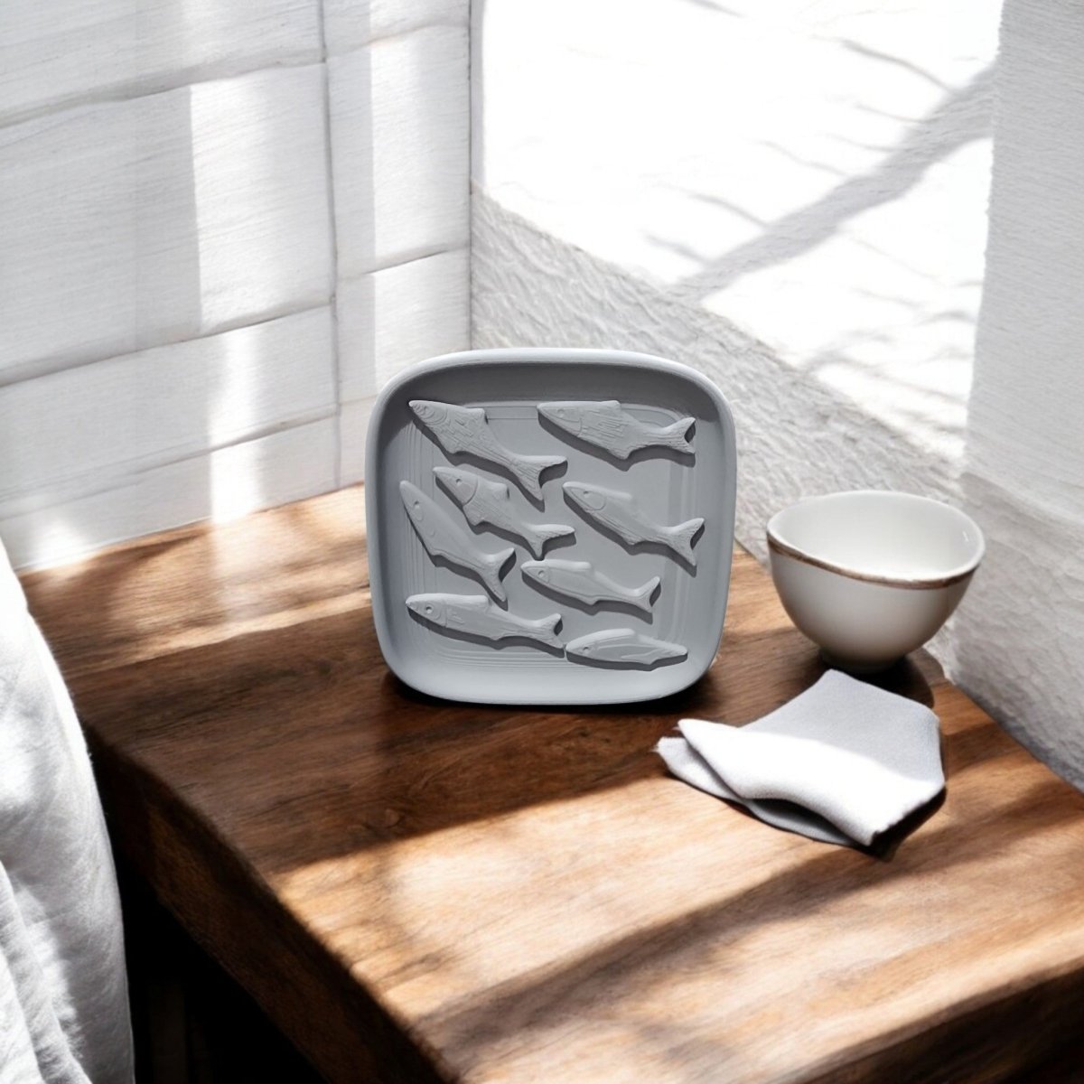 The Mirabilia Boutique Coastal Fish Decorative Tray, crafted with a gray silicone fish-shaped mold, rests on a wooden table beside a white bowl and folded napkin as sunlight casts shadows over the stylish ocean-inspired decor.