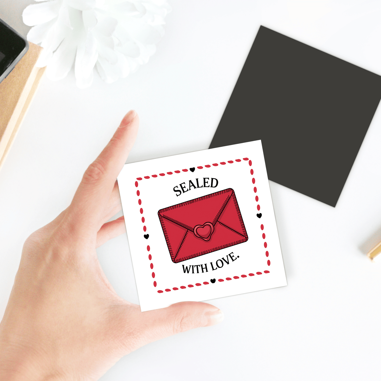 A hand holds Mirabilia Boutique's Sealed with Love Envelope Magnet, a square card design with a red envelope sealed by a heart. The romantic scene is completed by a white flower and black square paper.