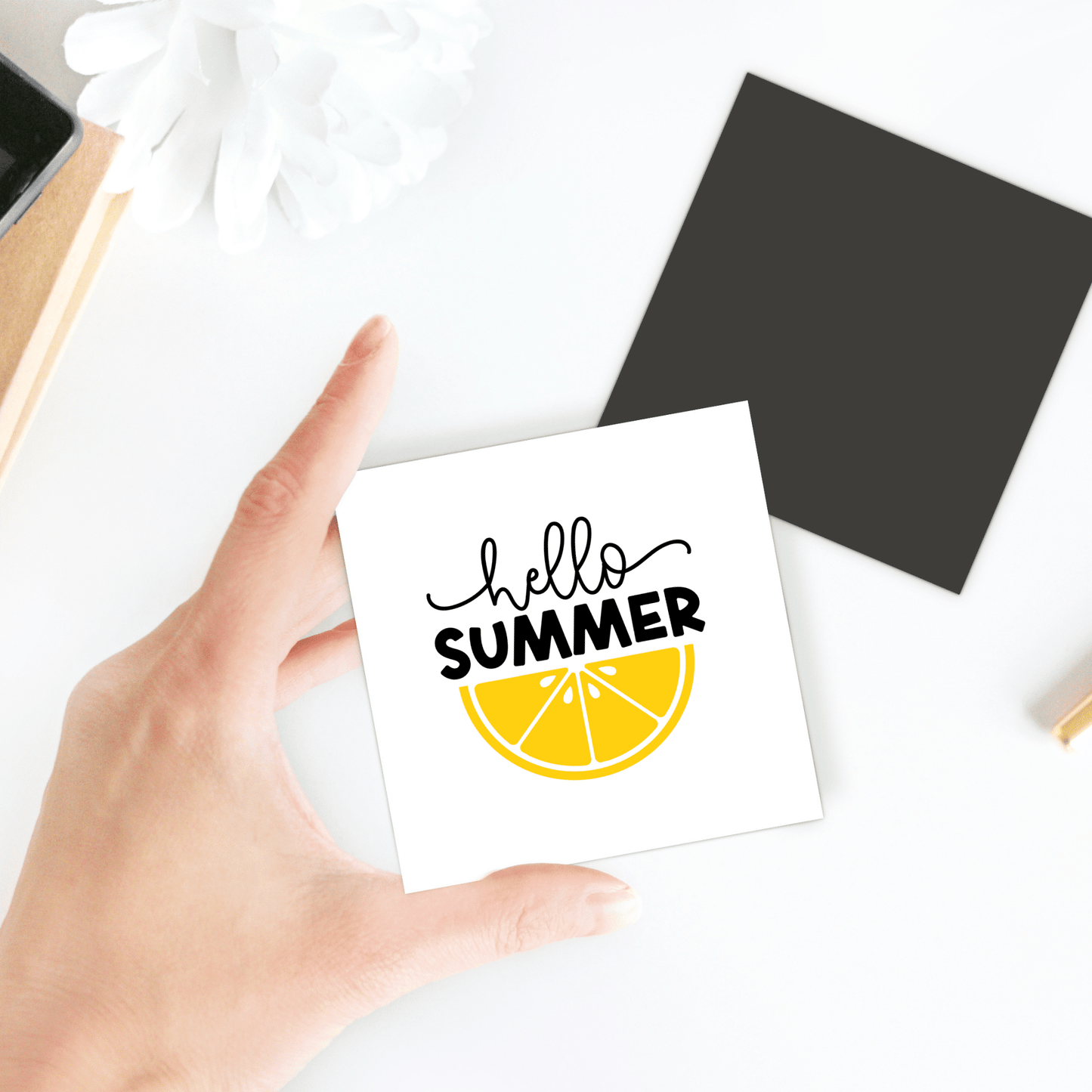 A hand holds the Hello Summer Lemon Magnet – Bold Citrus Fridge Art by Mirabilia Boutique, featuring "hello SUMMER" and a lemon slice. In the background, a black square and a white flower sit on a white surface.