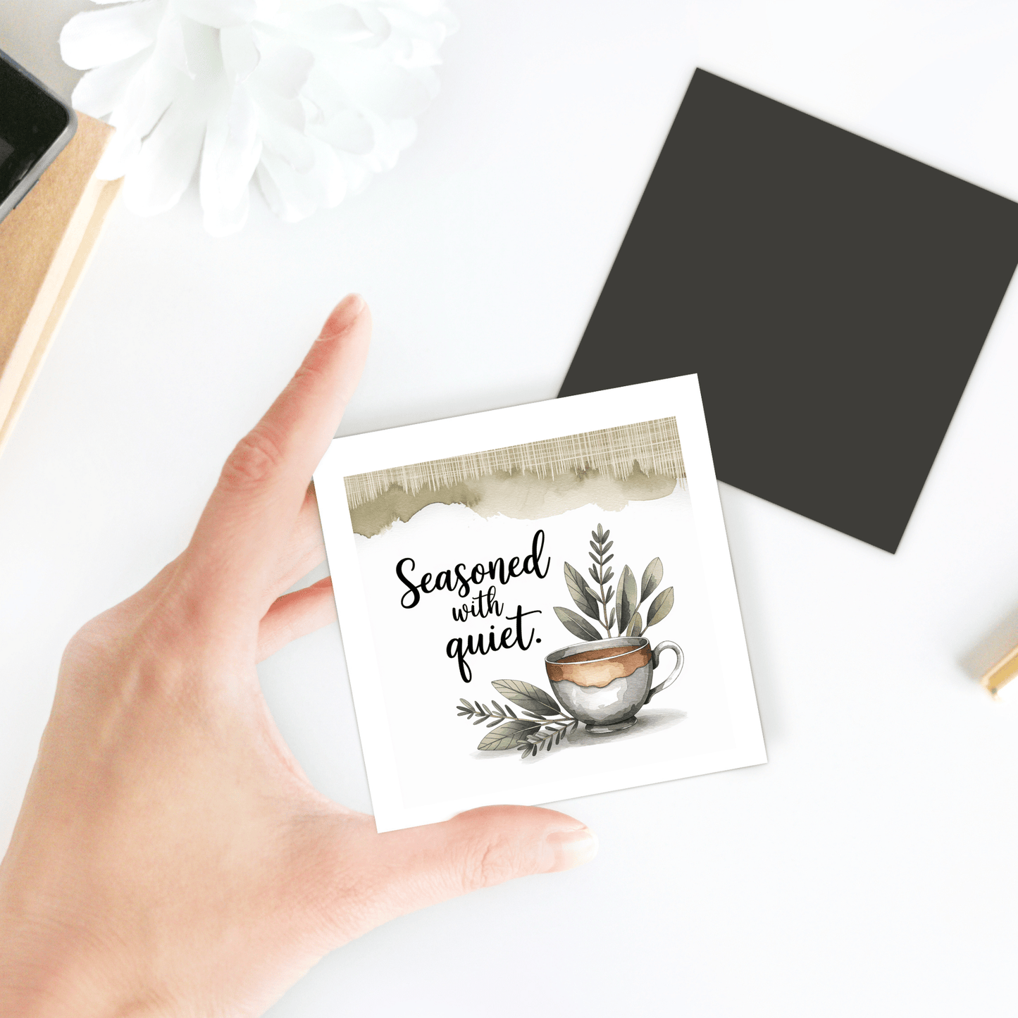 A hand holds the Mirabilia Boutique "Seasoned with Quiet Magnet | Calm Kitchen Decor Gift," featuring a coffee cup and green leaves—perfect for mindful spaces. A black card and white flower in the background create a serene vibe.
