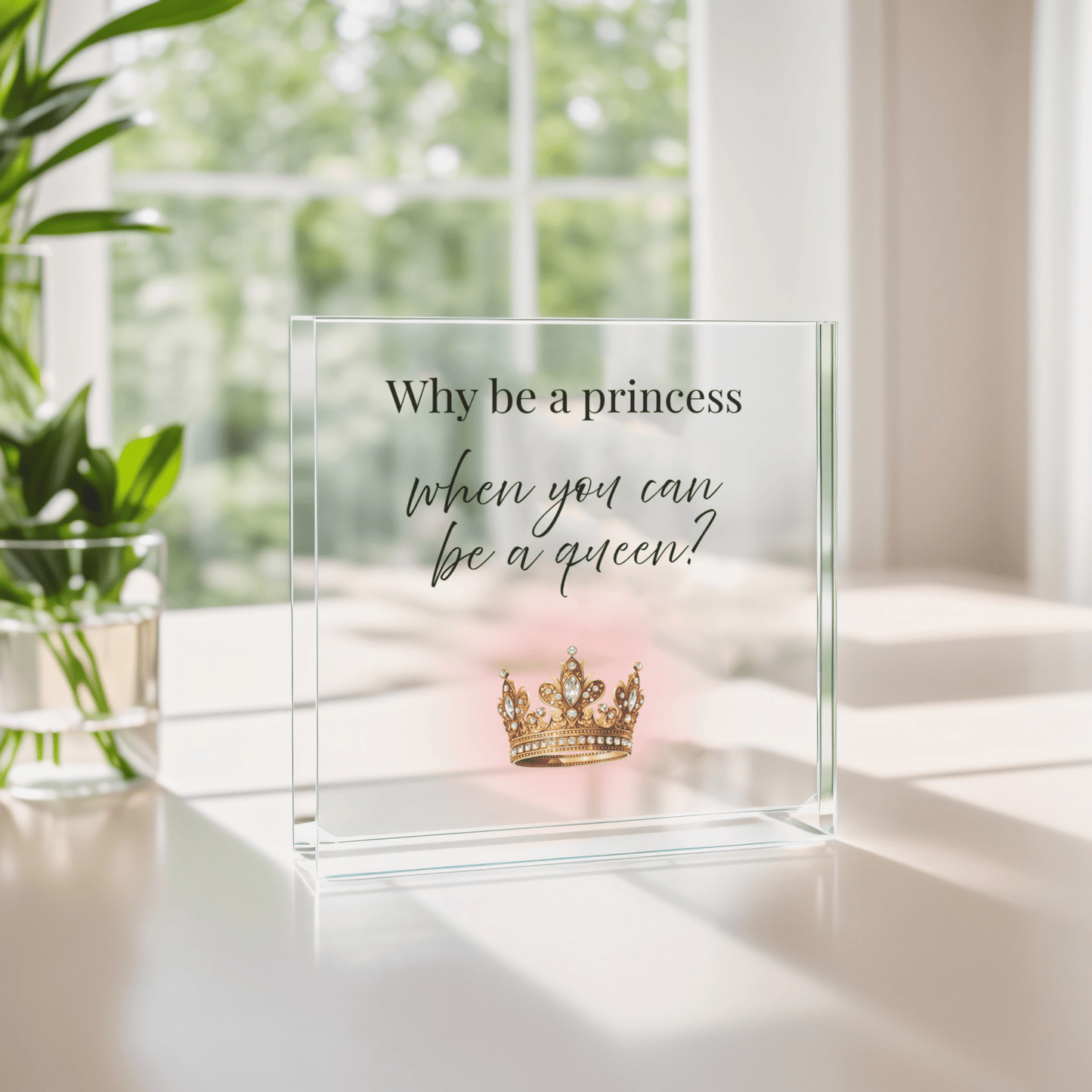 The Why Be a Princess When You Can Be a Queen Acrylic Block Gift for Her by Mirabilia Boutique features a gold crown and inspiring quote—an empowering decor piece, perfect for gifting or displaying among plants on a sunny windowsill.