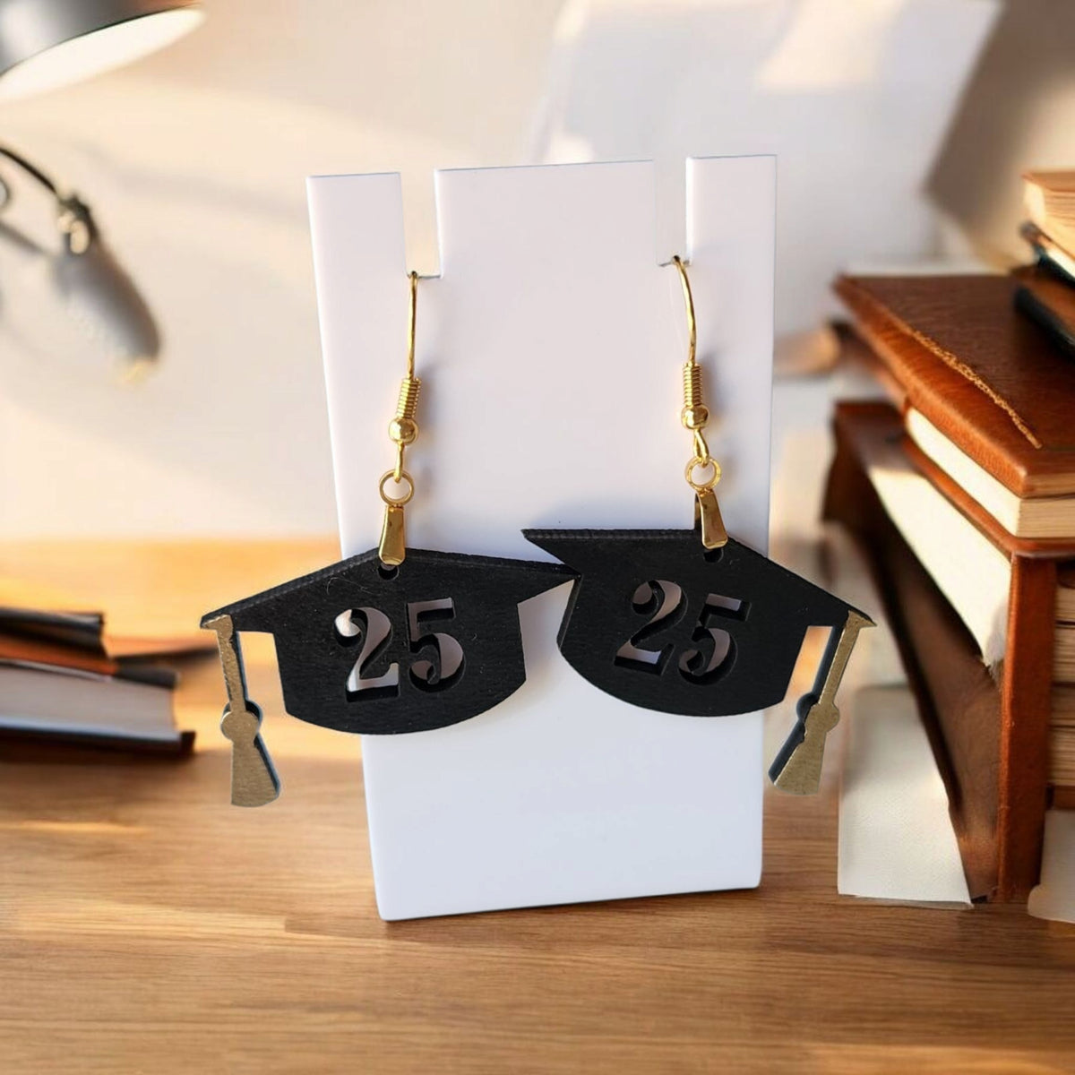Mirabilia Boutique’s Graduation Cap Earrings Class of 2025 feature black caps with “25” and gold hooks, hanging on a white stand against books and a lamp. Laser cut, they’re perfect grad party accessories or gifts for her.