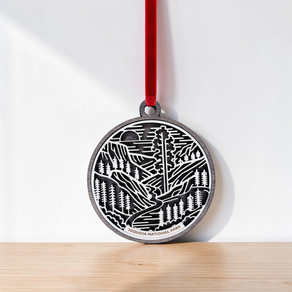 A round metal Sequoia National Park Line Art Christmas Ornament by Mirabilia Boutique, featuring a forest and mountain design and labeled "Sequoia National Park," hangs by a red ribbon against a white wall and rests on a light wooden surface—an ideal choice for anyone who loves giant sequoia art.
