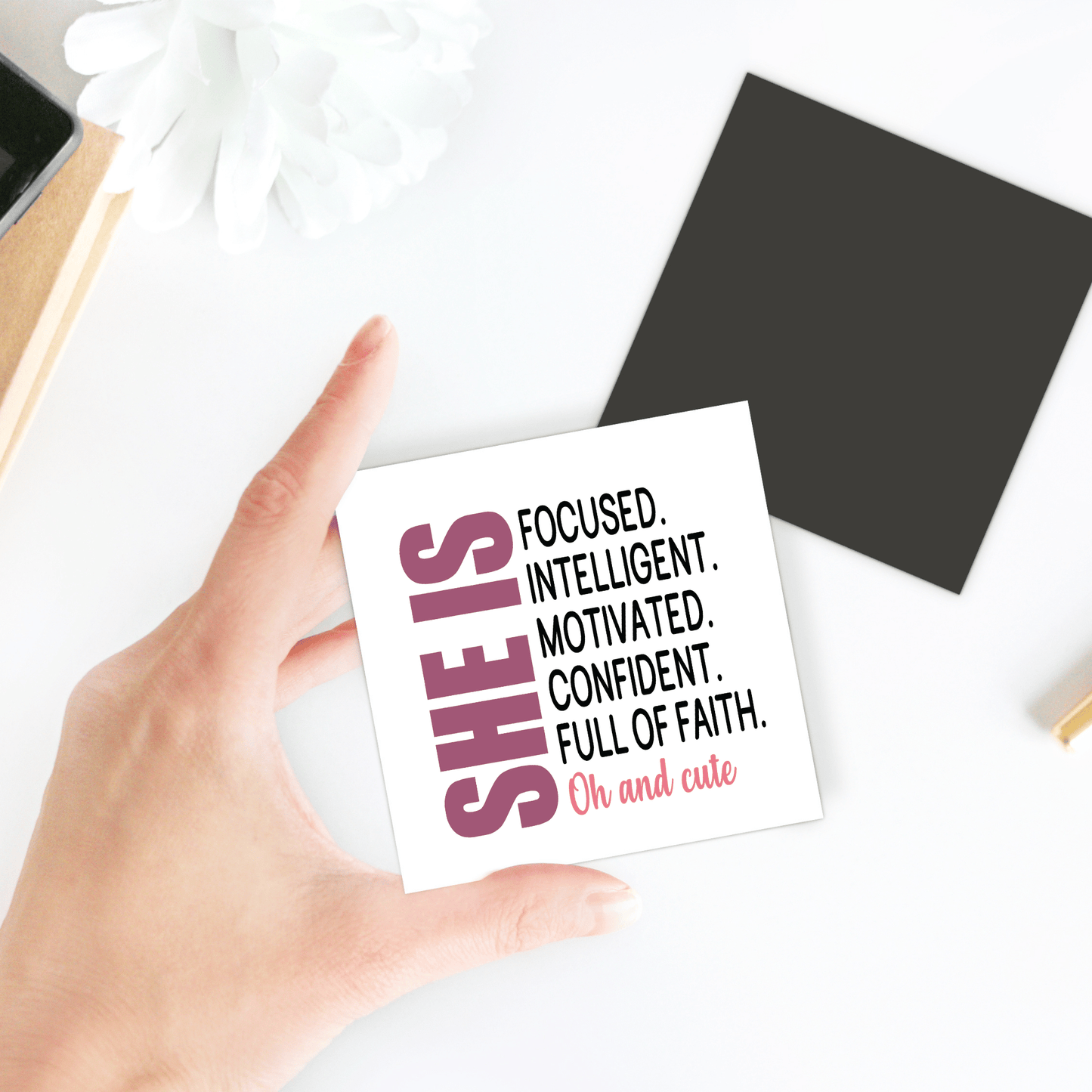 A hand holds the Mirabilia Boutique “She Is Motivated & Confident” magnet, reading: "SHE IS FOCUSED. INTELLIGENT. MOTIVATED. CONFIDENT. FULL OF FAITH. Oh and cute." An inspiring gift for her, featuring a black square and white flower on a white surface.