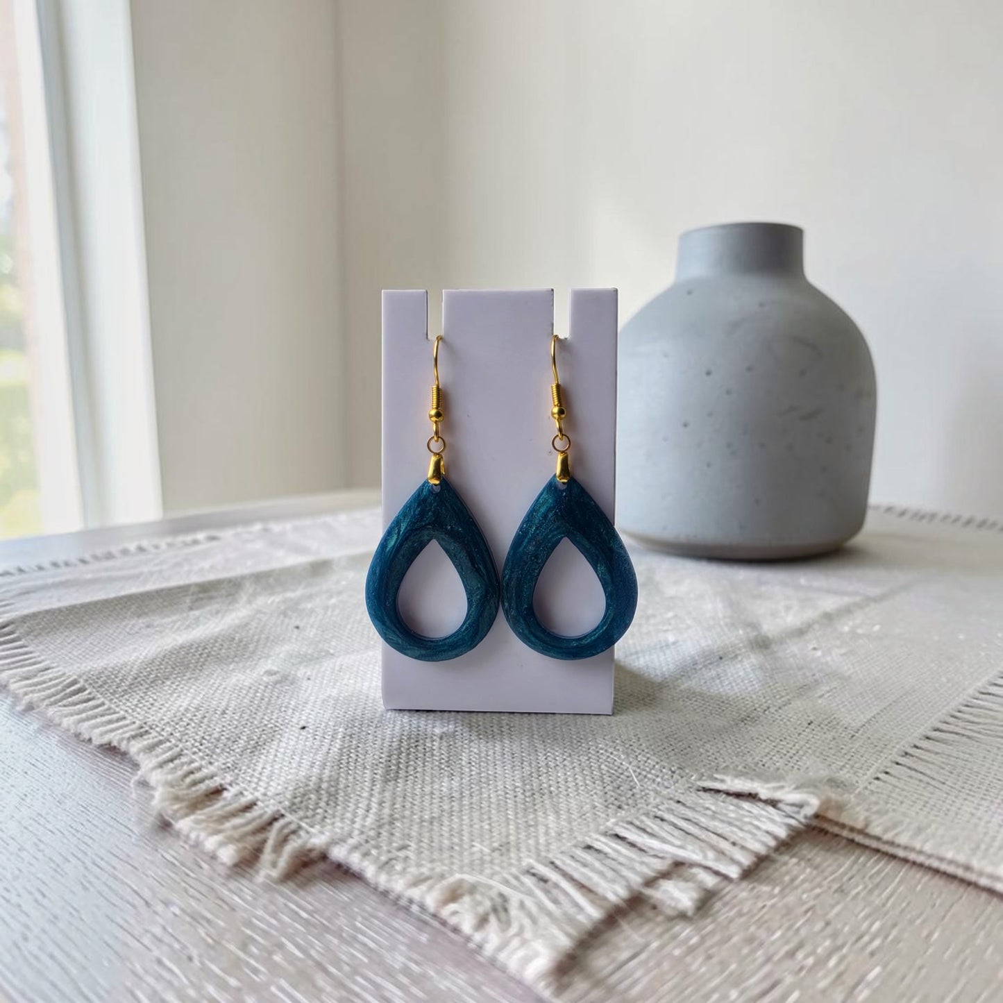 Mirabilia Boutique’s Mermaid Dust Teardrop Earrings, featuring blue drops and gold hooks on a white card stand, rest on a woven placemat with a gray vase behind—ideal for those who love mermaid-inspired jewelry.