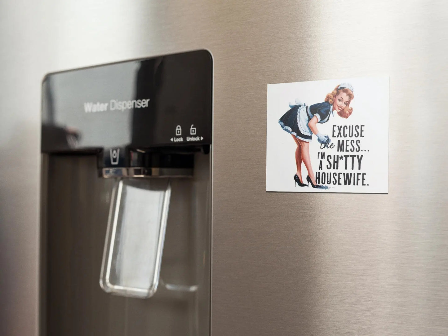 The stainless steel refrigerator with a built-in water dispenser is adorned with the Retro "Excuse the Mess" Shitty Housewife Magnet from Mirabilia Boutique, adding humorous fridge decor featuring a woman declaring, "Excuse the mess... I'm a shitty housewife." This quirky kitchen accessory brings charm and laughter to your culinary space.