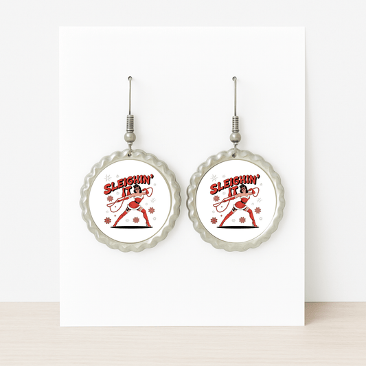 The Sleighin' It Earrings by Mirabilia Boutique are round, silver holiday earrings featuring a dancing Santa and "Sleighin' It" text in red—fun Christmas accessories for women, shown on a white background.