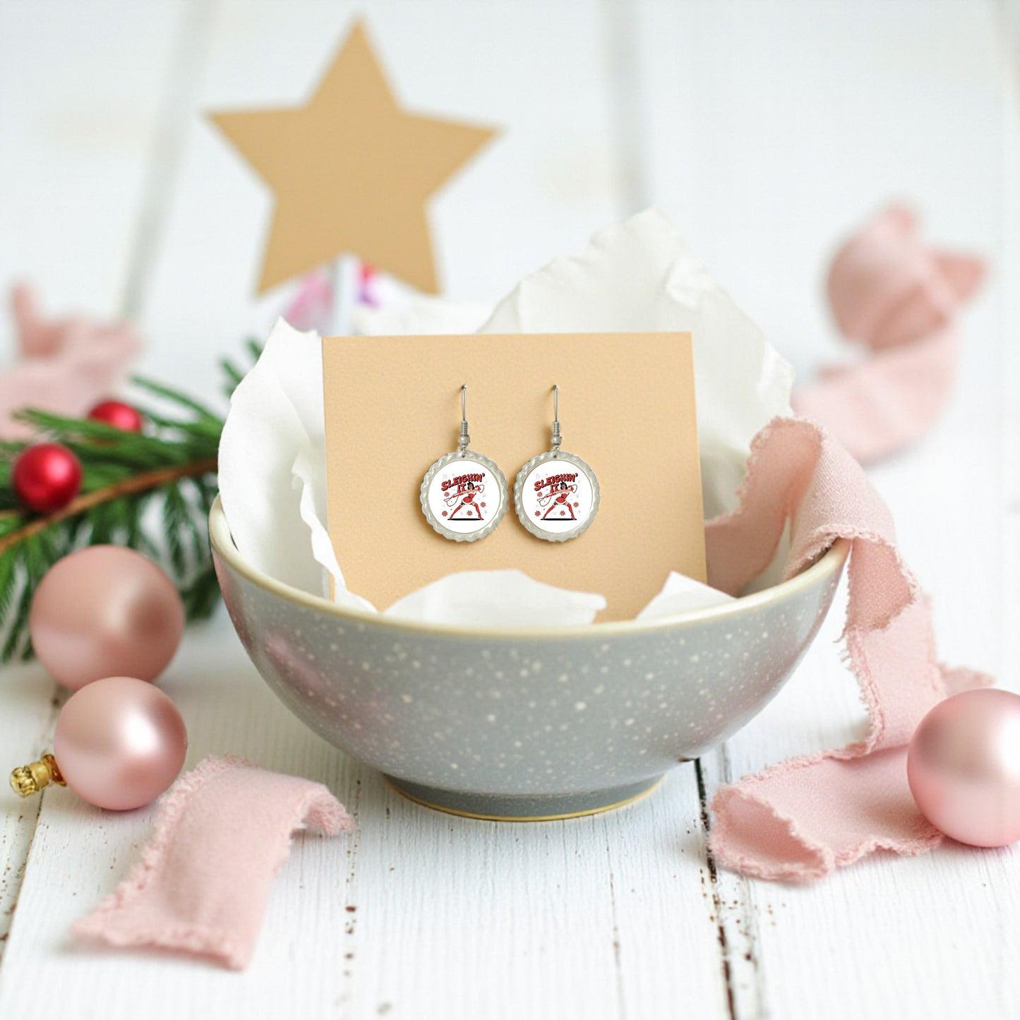 The Sleighin' It Earrings by Mirabilia Boutique are festive round holiday earrings displayed in a gray bowl with tissue paper, pink ornaments, ribbon, pine, and a gold star—perfect Christmas party accessories.