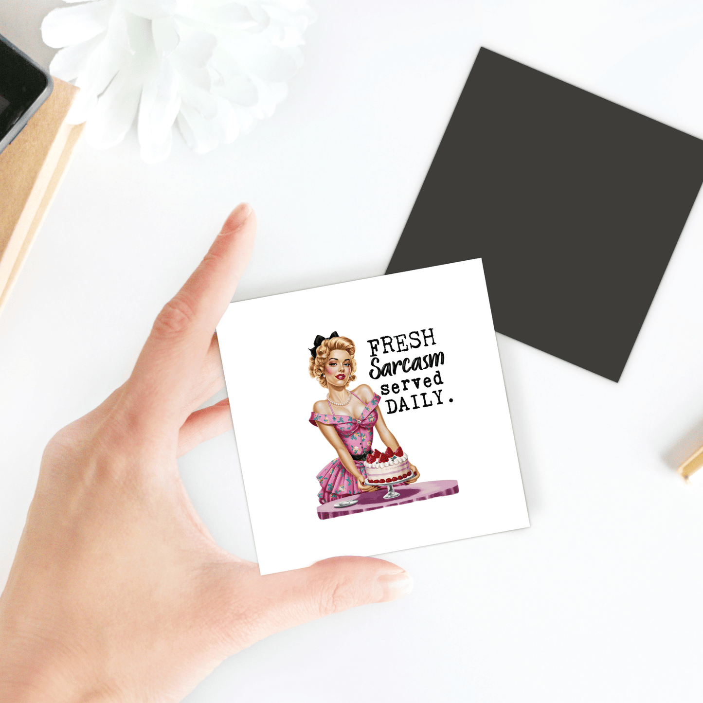 A hand displays the Mirabilia Boutique "Fresh Sarcasm Served Daily" 3x3 retro UV printed magnet—featuring a witty woman with a cake—ideal as sassy fridge or kitchen décor and a funny gift for sarcasm lovers.