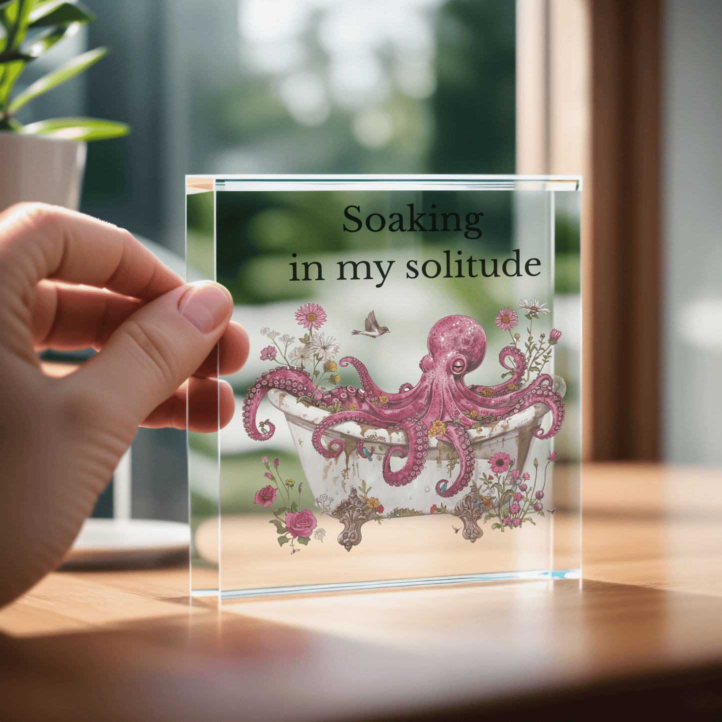 A hand displays the Soaking in My Solitude Art Block by Mirabilia Boutique, showing a pink octopus relaxing in a floral tub with "Soaking in my solitude," ideal as quirky decor or a solitude gift.