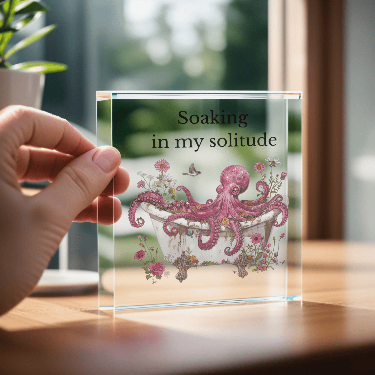 A hand holds the Soaking in My Solitude Art Block by Mirabilia Boutique, featuring a pink octopus in a floral bathtub. Sunlight streams through a window, making this whimsical decor the perfect solitude gift for any space.