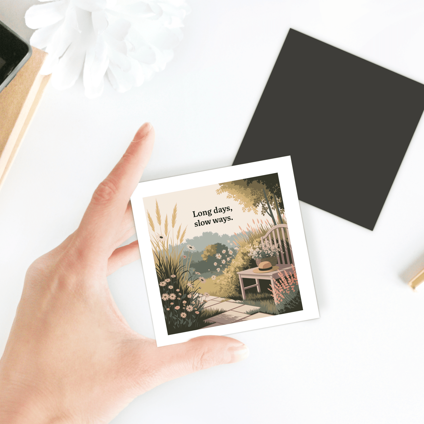 A hand holds the "Long Days, Slow Ways Magnet – Peaceful Summer Garden Quote" by Mirabilia Boutique, showing a garden bench, flowers, and teacup. Perfect as a mindfulness gift or summer decor. Nearby are a black card and white flower on a white surface.