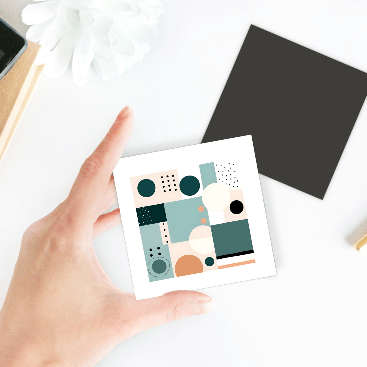 A hand holds the Coastal Geometry Mid Century Modern Magnet by Mirabilia Boutique, featuring an abstract geometric design in muted tones. In the background, a black square and a white flower rest on a white surface.