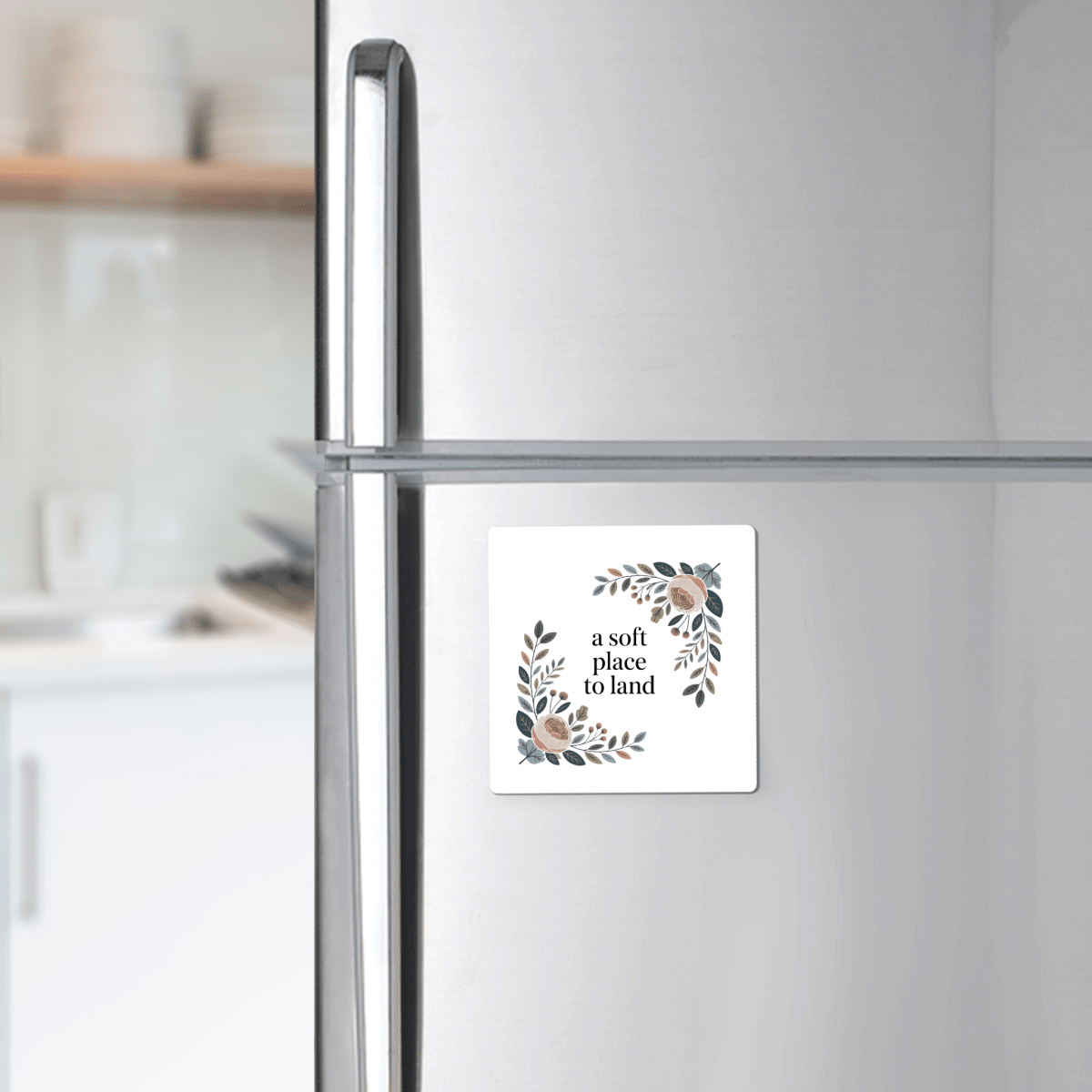 A silver fridge displays the Mirabilia Boutique "A Soft Place to Land Floral Magnet," a square cottagecore piece with a floral wreath and uplifting words. Blurred shelves and kitchen counter appear in the background.