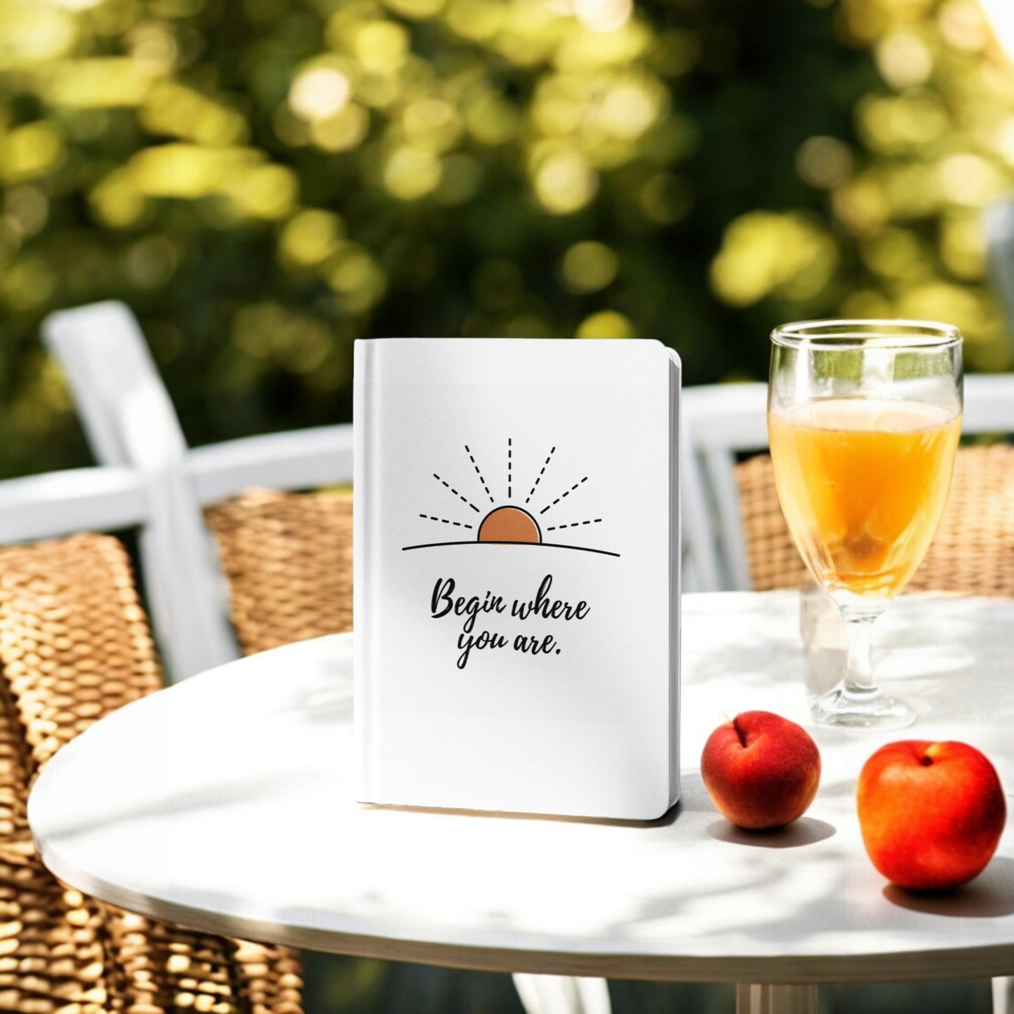 The Begin Where You Are Journal – Sunrise Quote Notebook by Mirabilia Boutique, adorned with a sun and an inspiring quote, sits outdoors beside juice and peaches—ideal for daily reflections.