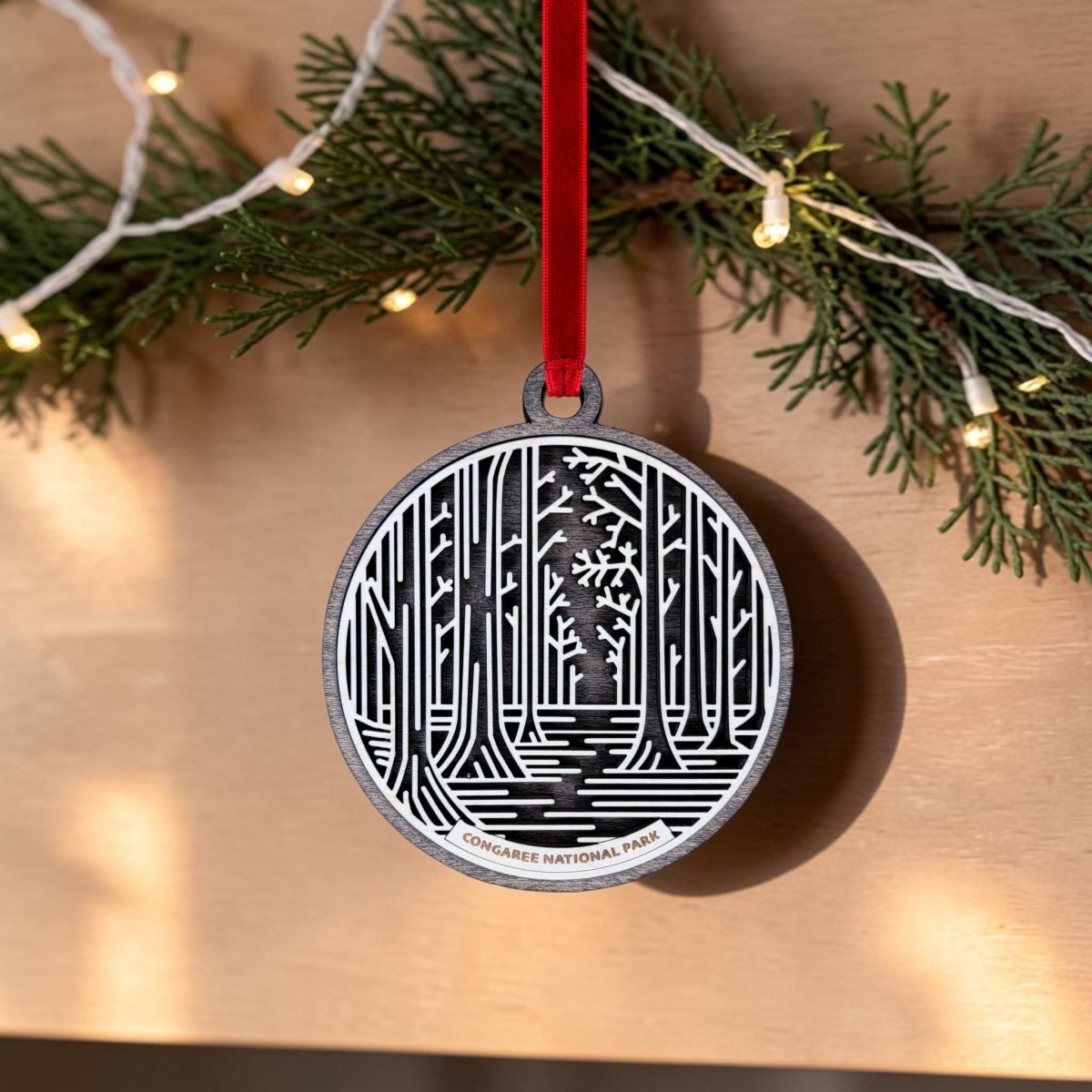 A round handcrafted wooden ornament from Mirabilia Boutique, featuring a black and white forest illustration and labeled "Congaree National Park," hangs from a red ribbon amidst greenery and string lights—the perfect Christmas keepsake for old-growth forest enthusiasts.