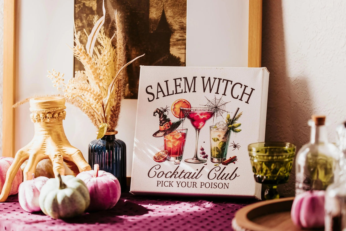 The Mirabilia Boutique Salem Witch Cocktail Club Halloween Canvas (8x8) is styled on a festive bar cart with pumpkins, a gold hand statue, glassware, and dried flowers—perfect spooky decor in warm sunlight.