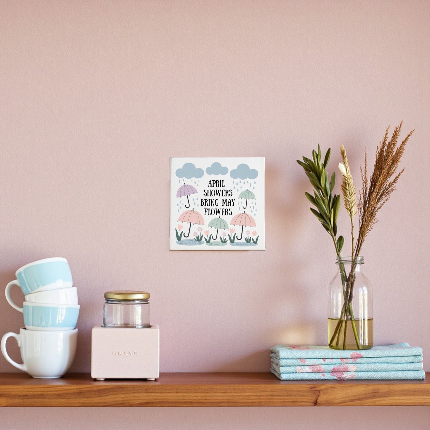 A wooden shelf holds blue cups, a glass jar, a beige box, dried flowers in a vase, and folded towels. Above is the Mirabilia Boutique "April Showers Bring May Flowers" Canvas Art with cheerful spring motifs—perfect for any nursery.