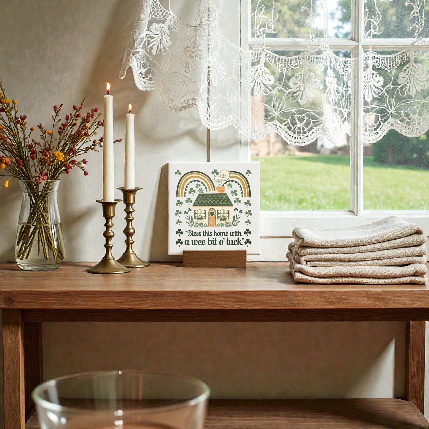 A wooden table holds stacked towels, two lit candles, a vase with dried flowers, and Mirabilia Boutique’s "A Wee Bit of Luck Canvas Sign – Irish Cottage Charm" near a lace-curtained window overlooking a green yard.