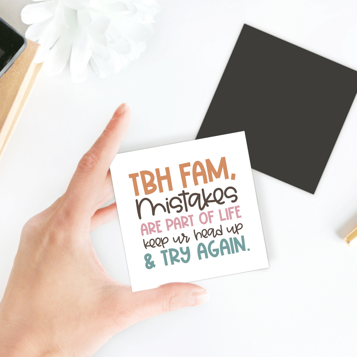 A hand holds the Mirabilia Boutique "Growth Mindset Teacher Magnet | Try Again Quote." With a white flower, black square, and gold pen on the desk, it’s perfect for motivational classroom decor or a thoughtful teacher appreciation gift.