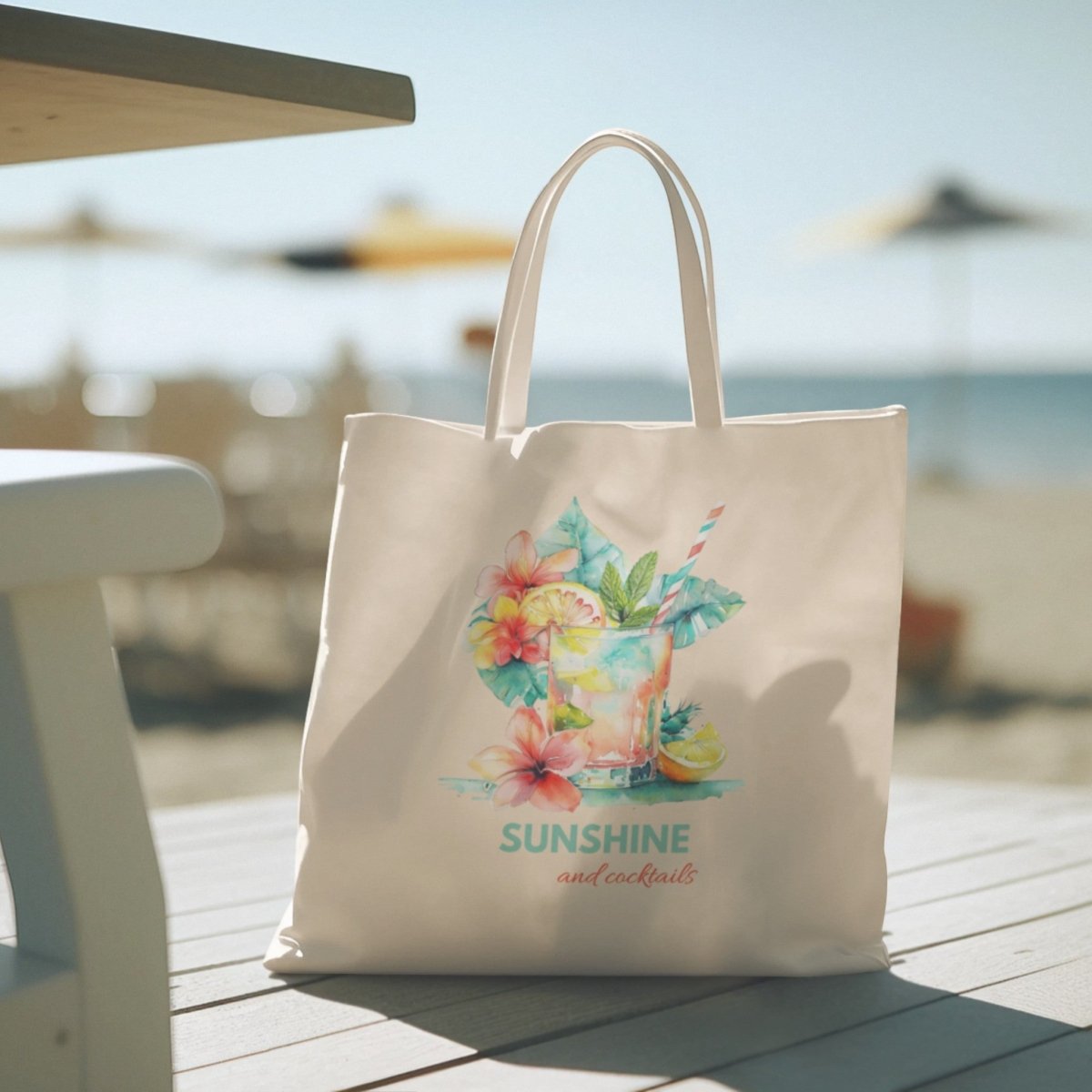 The Mirabilia Boutique "Sunshine and Cocktails Canvas Tote Bag"—featuring a vibrant beach drink, floral artwork, and the phrase "Sunshine and cocktails" on white canvas—rests on a table at a sunny beach, with chairs and umbrellas softly blurred in the background.