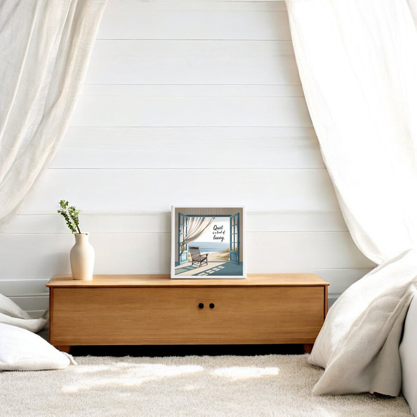 A minimalist bedroom highlights Mirabilia Boutique’s “Quiet Is a Kind of Luxury Canvas – Peaceful Summer Wall Art” above a low bench, paired with white curtains and green foliage—an elegant touch of quiet luxury decor.