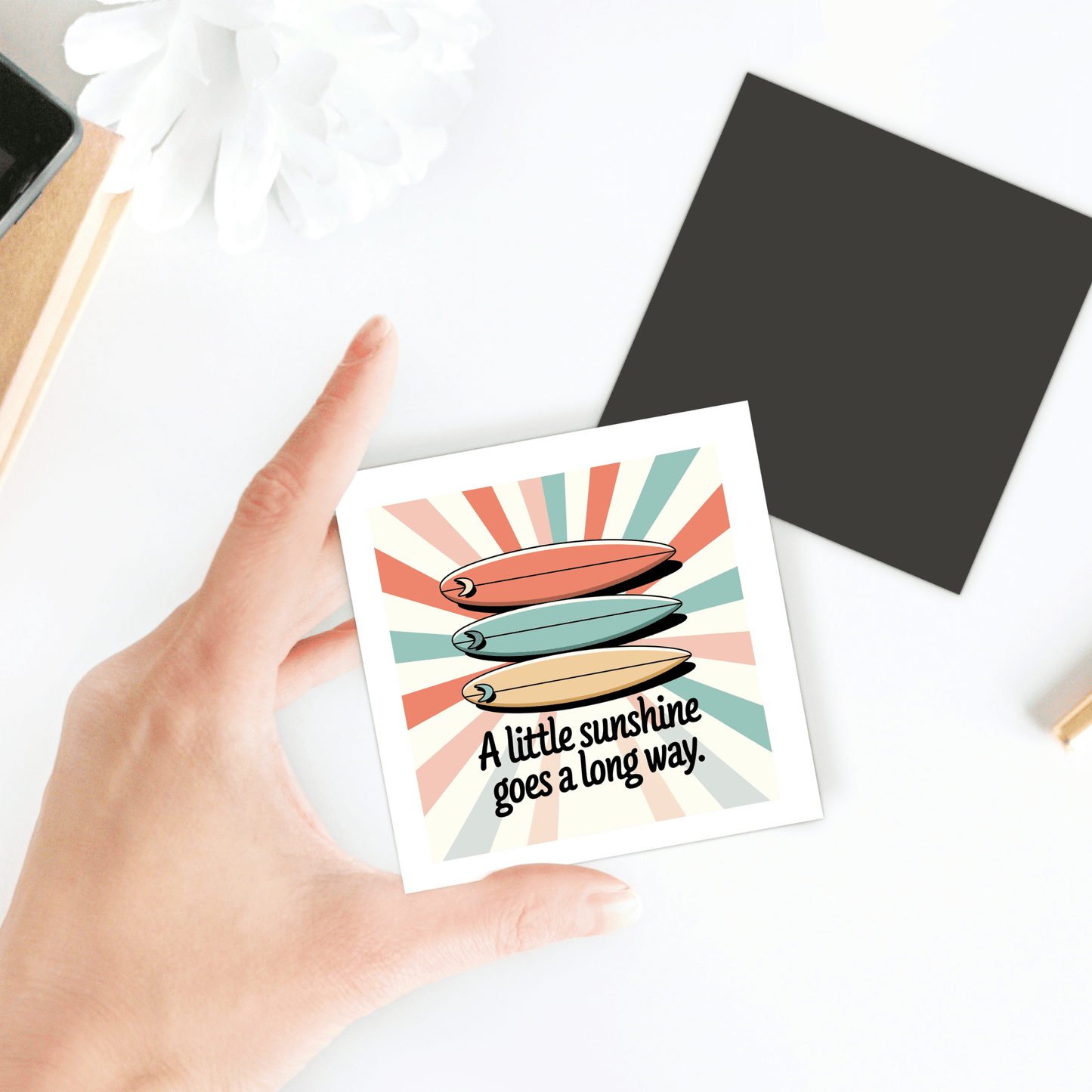 A hand holds the "A Little Sunshine Magnet – Retro Surf & Sun Vibes" by Mirabilia Boutique featuring two surfboards and sunny text, shown with sunshine-themed decor, a white flower, gold pen, and black square on a bright surface.