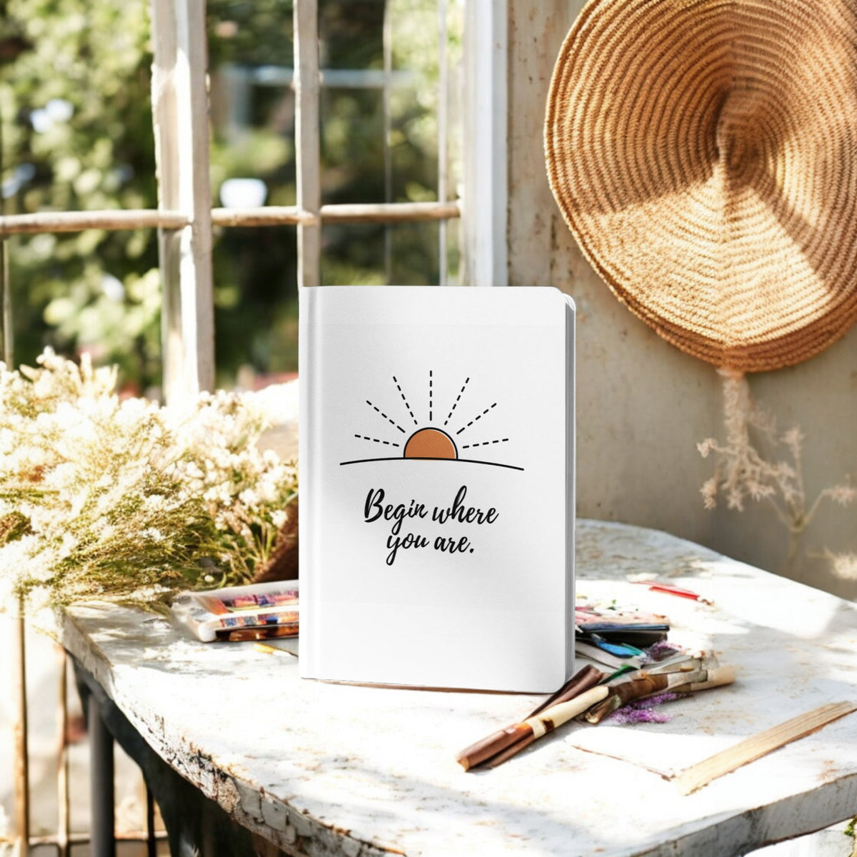 The Begin Where You Are Journal – Sunrise Quote Notebook by Mirabilia Boutique sits on a sunlit table with art supplies, dried flowers, and a woven hat nearby—perfect for daily writing amid greenery outside the window.