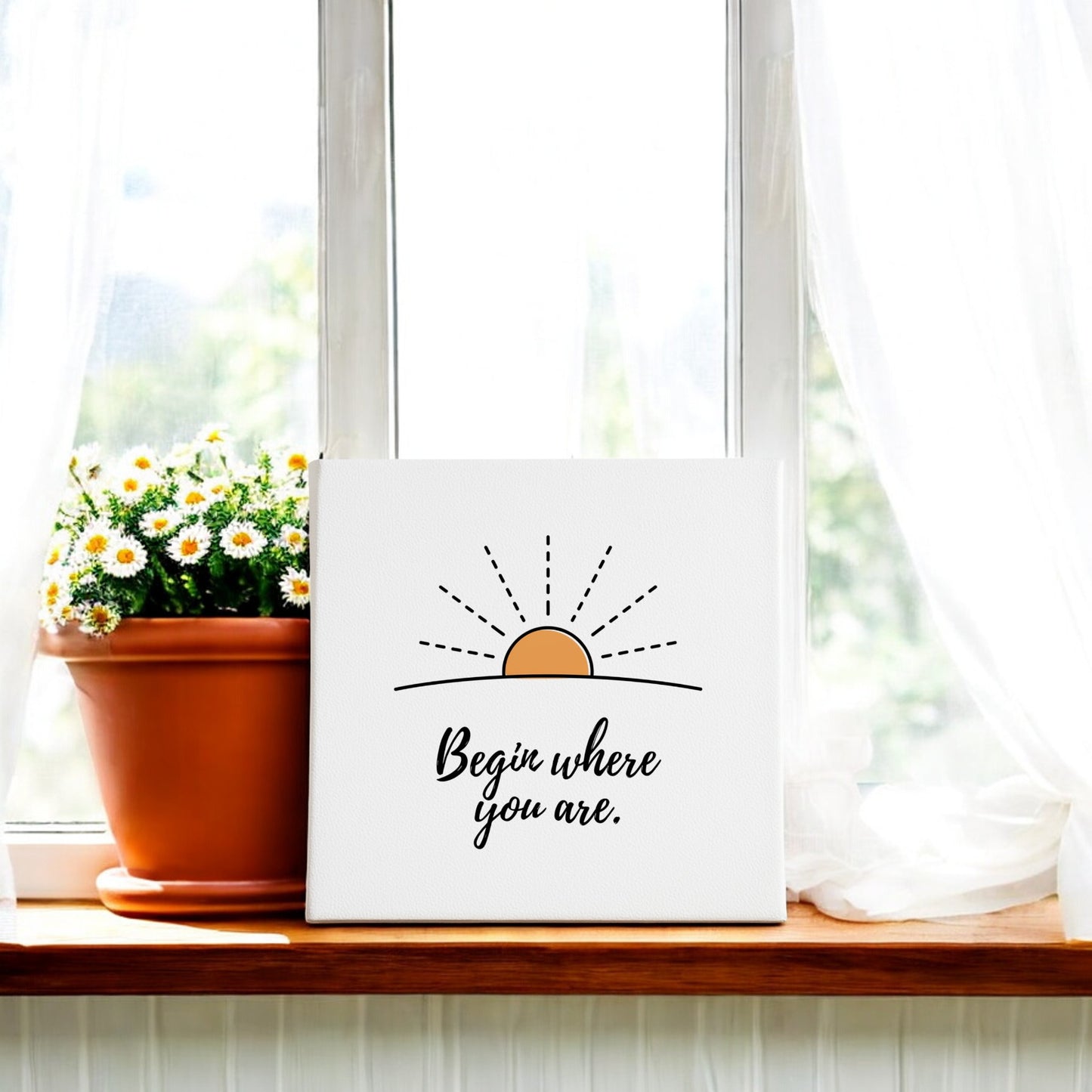 A potted daisy sits on a sunny windowsill beside the Mirabilia Boutique "Begin Where You Are Canvas – Sunrise Wall Affirmation," framed by white curtains as sunlight highlights this motivational artwork.