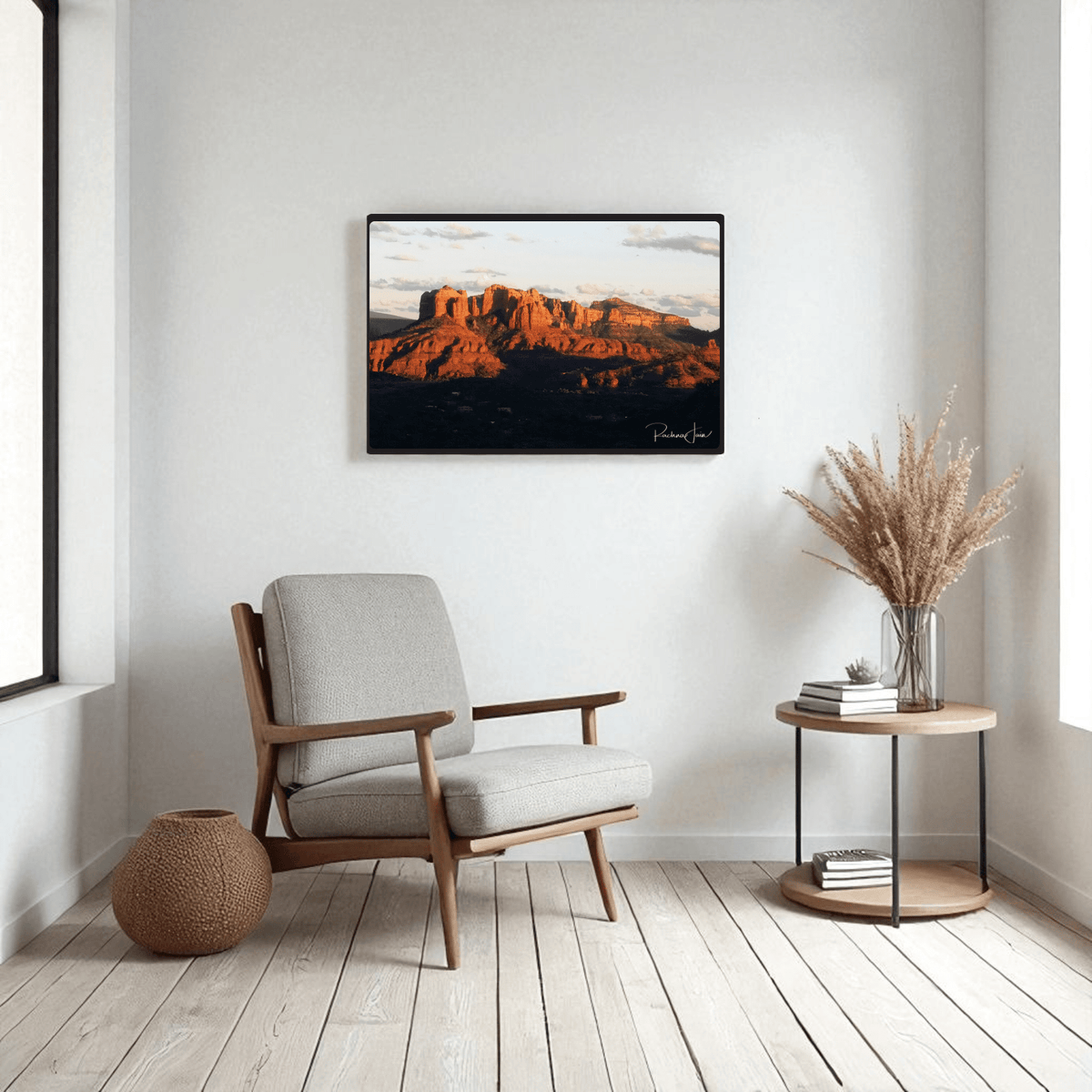A modern, minimalist living room with a gray armchair, round side table holding a vase of dried grass, woven basket, and Mirabilia Boutique’s "Sunset in Sedona" framed art print by Rachna Jain on white walls with light wood floors.