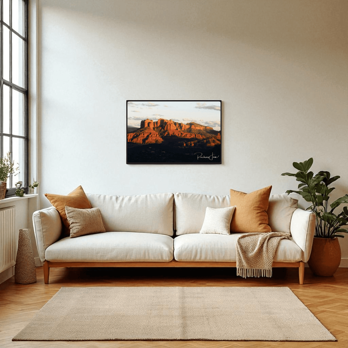 A bright living room features a white sofa, cozy pillows, potted plants, and a large window. Above the sofa hangs "Sunset in Sedona" framed art print by Rachna Jain from Mirabilia Boutique—a stunning southwest landscape.