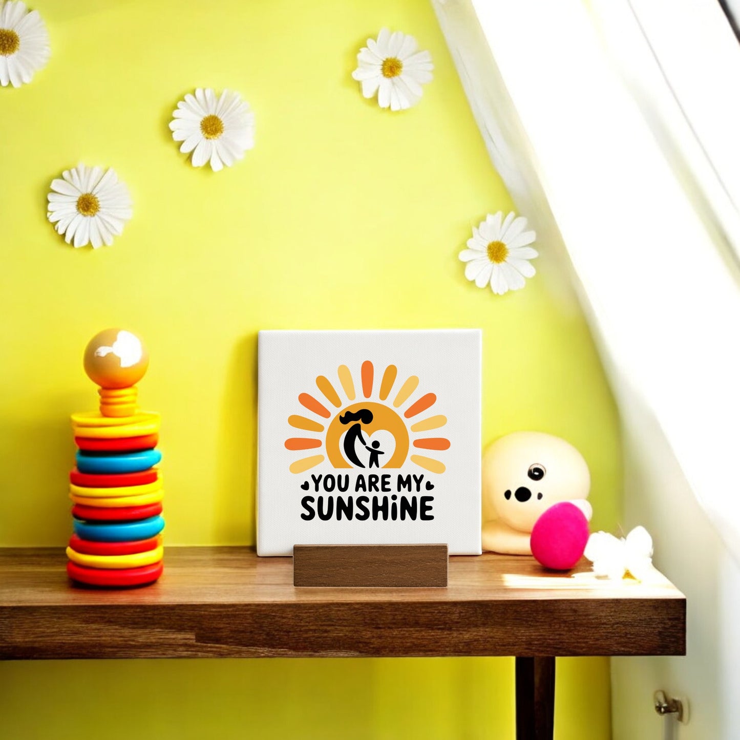 A wooden table displays the “You Are My Sunshine Mini Sign – Mom & Child Canvas” by Mirabilia Boutique, a colorful stacking toy, and a white decor piece. Sunlight and daisy accents create an uplifting Mother’s Day gift scene.
