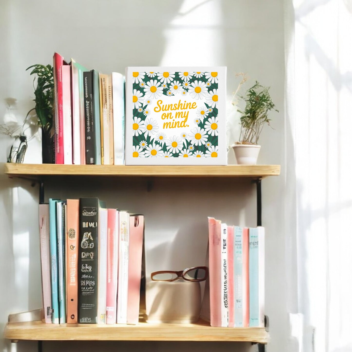 A wooden bookshelf displays colorful books, glasses, potted plants, and Mirabilia Boutique’s “Sunshine on My Mind” 8x8 Daisy Wall Art Canvas. Sunlight streams in, creating cheerful summer home decor vibes.