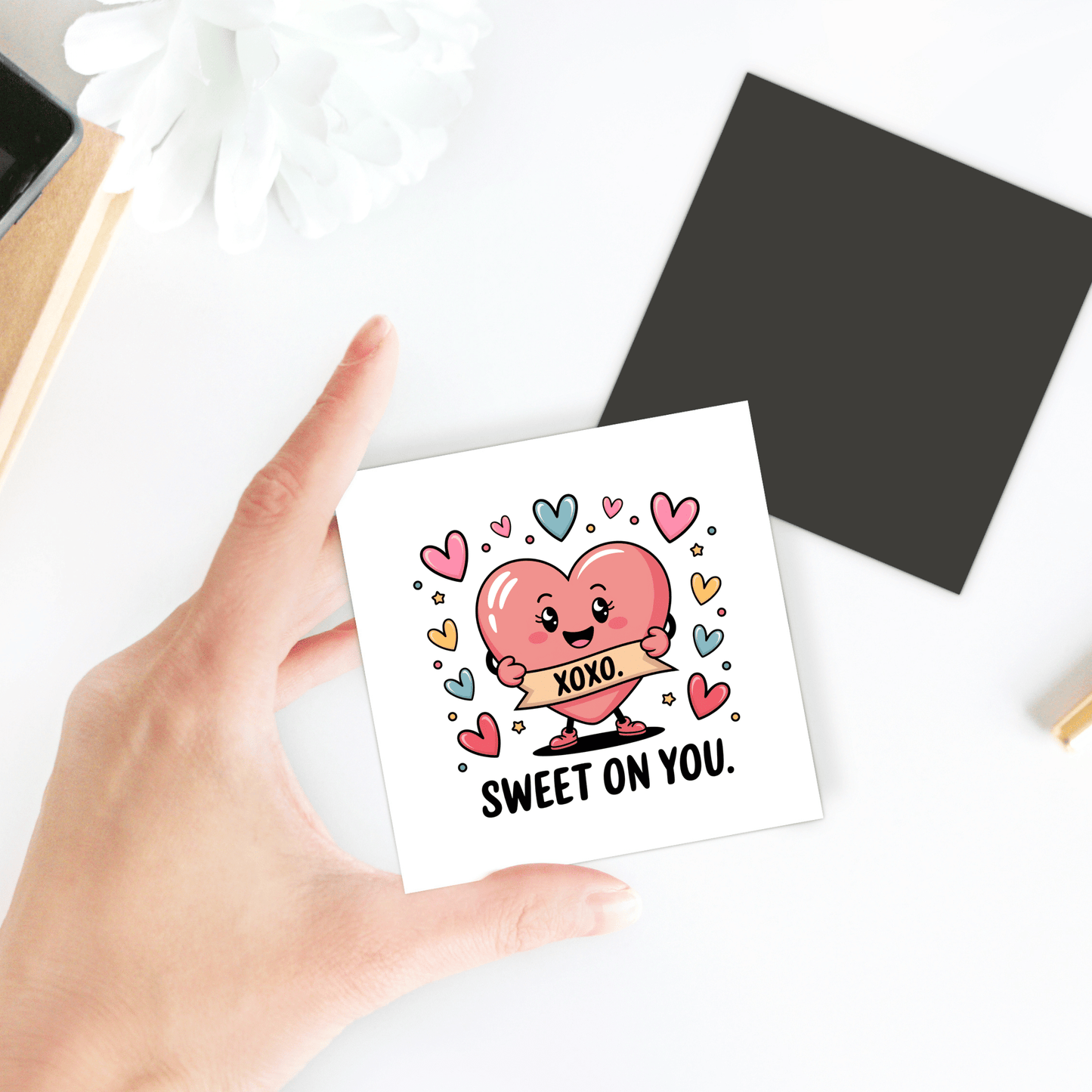 A hand holds the Sweet on You Valentine Magnet by Mirabilia Boutique—a square card with a pink heart character, colorful hearts, and an "XOXO" banner. Perfect as a romantic kitchen decor or love note. Flower and blank card accompany it on a white surface.