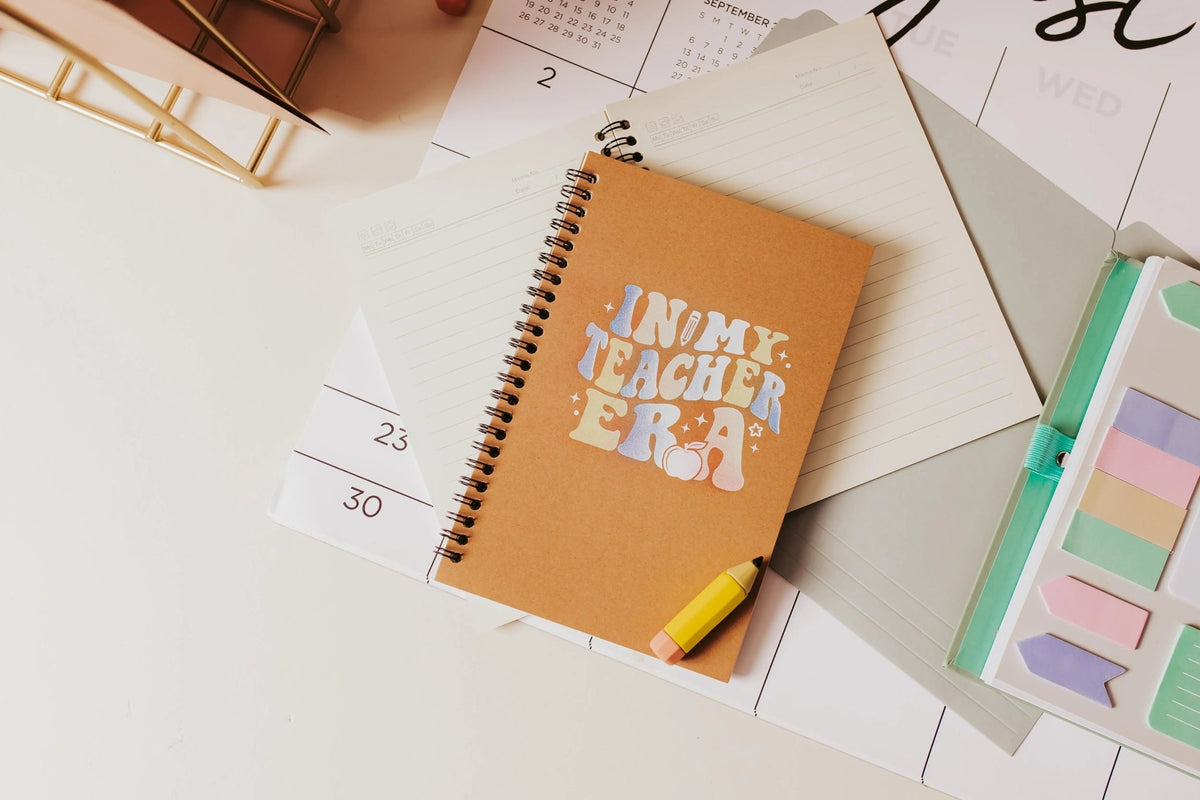 The "In My Teacher Era Notebook" by Mirabilia Boutique, an aesthetic spiral journal, sits on a calendar with lined paper, pastel sticky notes, and highlighters—the perfect back-to-school teacher gift.