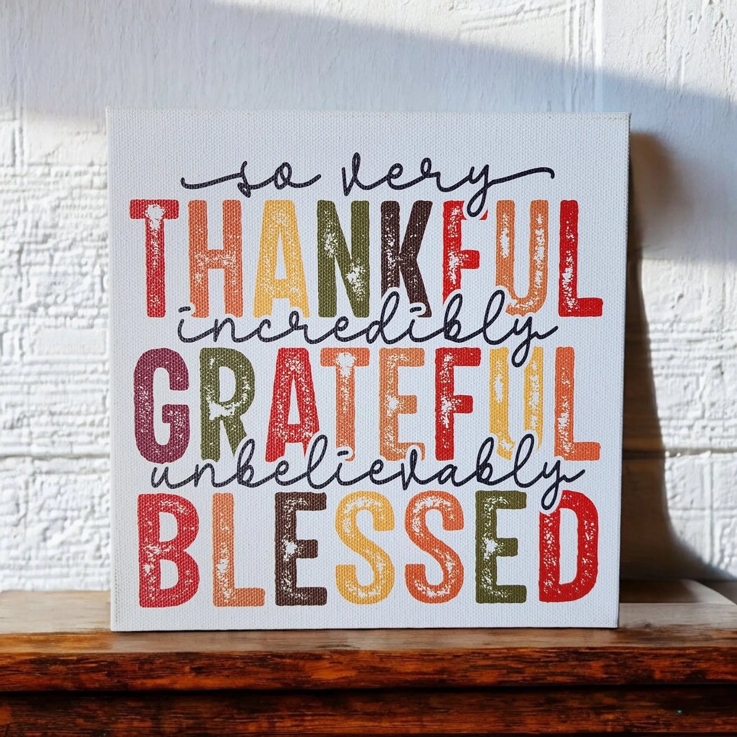 A canvas from Mirabilia Boutique, featuring colorful and bold text, is propped against a white textured wall. The "Thankful Grateful Blessed Inspirational Sign for Seasonal Home Decor" reads: "so very Thankful, incredibly Grateful, unbelievably Blessed." Embracing Thanksgiving colors—shades of red, orange, yellow, green, and brown—this home decor sign is perfect for any cozy space. The canvas rests gracefully on a wooden surface.