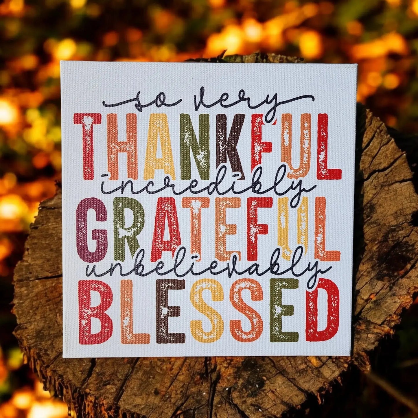 The Thankful Grateful Blessed Inspirational Sign for Seasonal Home Decor by Mirabilia Boutique features the words "So very thankful, incredibly grateful, unbelievably blessed" in capital letters. The text alternates autumn colors such as orange, red, green, and yellow with a background of fall leaves on a white canvas displayed on a tree stump, making it perfect home decor for showcasing Thanksgiving colors.