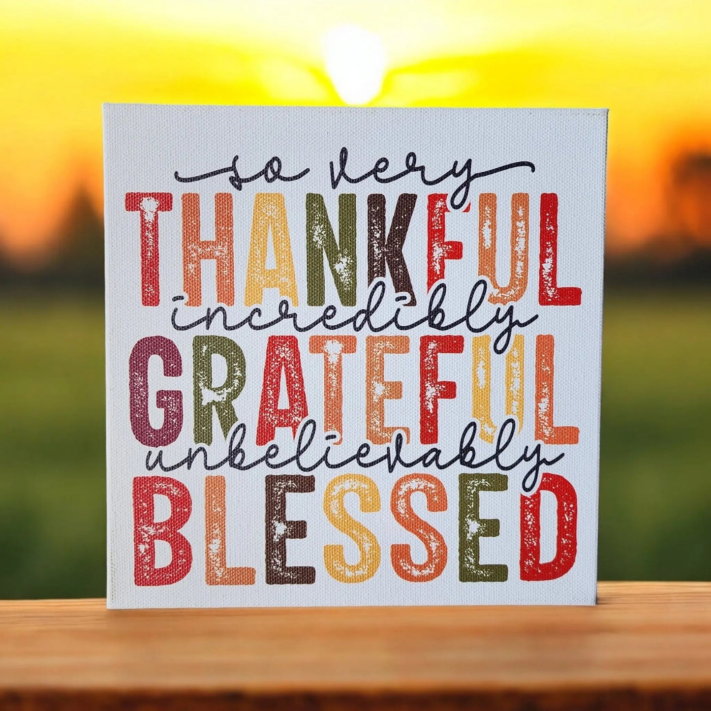 Introducing the "Thankful Grateful Blessed Inspirational Sign for Seasonal Home Decor" from Mirabilia Boutique, a square sign featuring the vibrant message "So very thankful, incredibly grateful, unbelievably blessed" in beautiful Thanksgiving colors and a variety of fonts. The backdrop showcases a softly focused sunrise or sunset scene, with the sun's radiant glow enhancing the charm of this piece. Perfectly designed to rest on any wooden surface, it adds warmth and inspiration to your home decor.