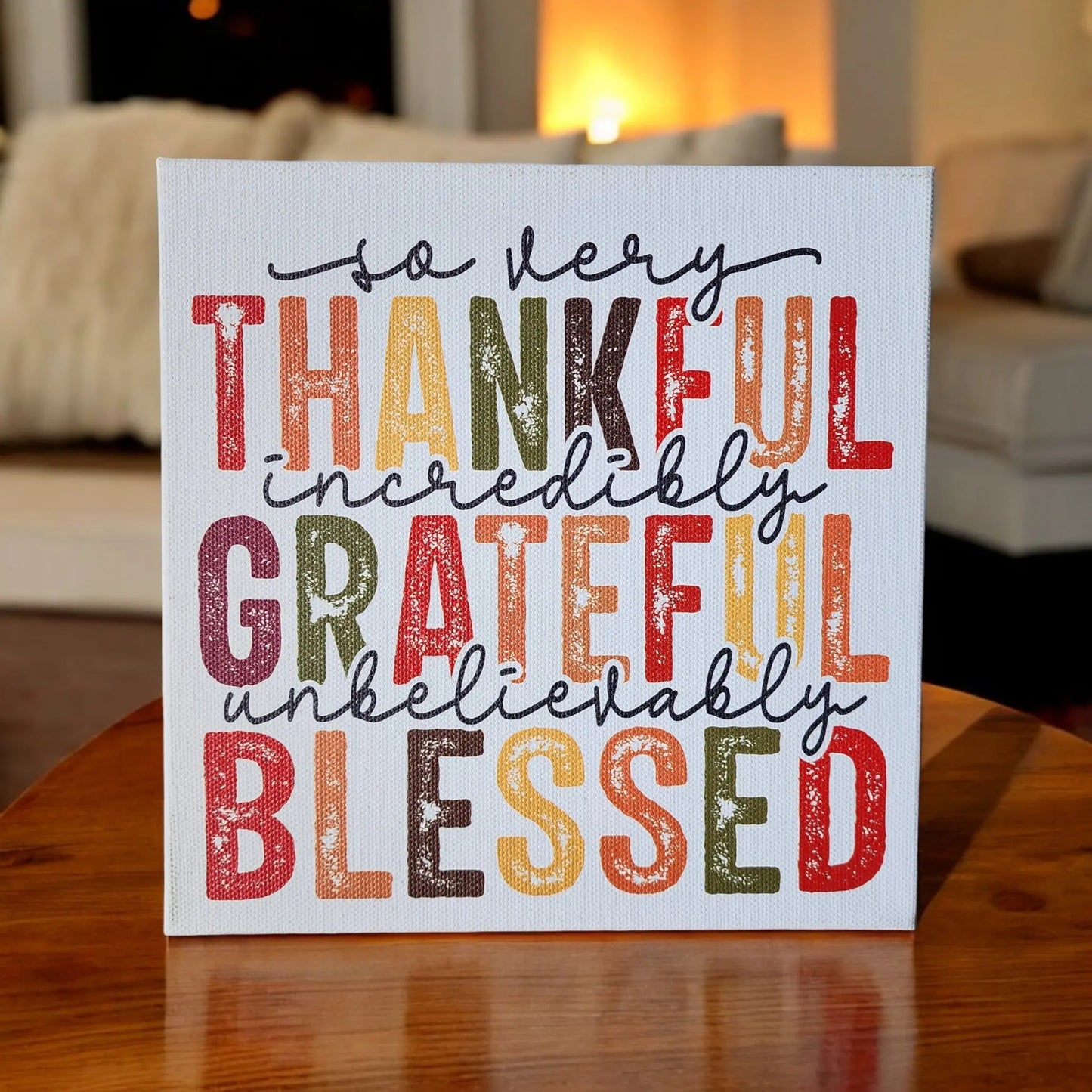 A square Thankful Grateful Blessed Inspirational Sign for Seasonal Home Decor by Mirabilia Boutique features decorative text on a wooden surface. The sign reads, "So very THANKFUL, incredibly GRATEFUL, unbelievably BLESSED" in Thanksgiving colors: orange, yellow, red, green, and brown. In the background, a cozy living room is visible.