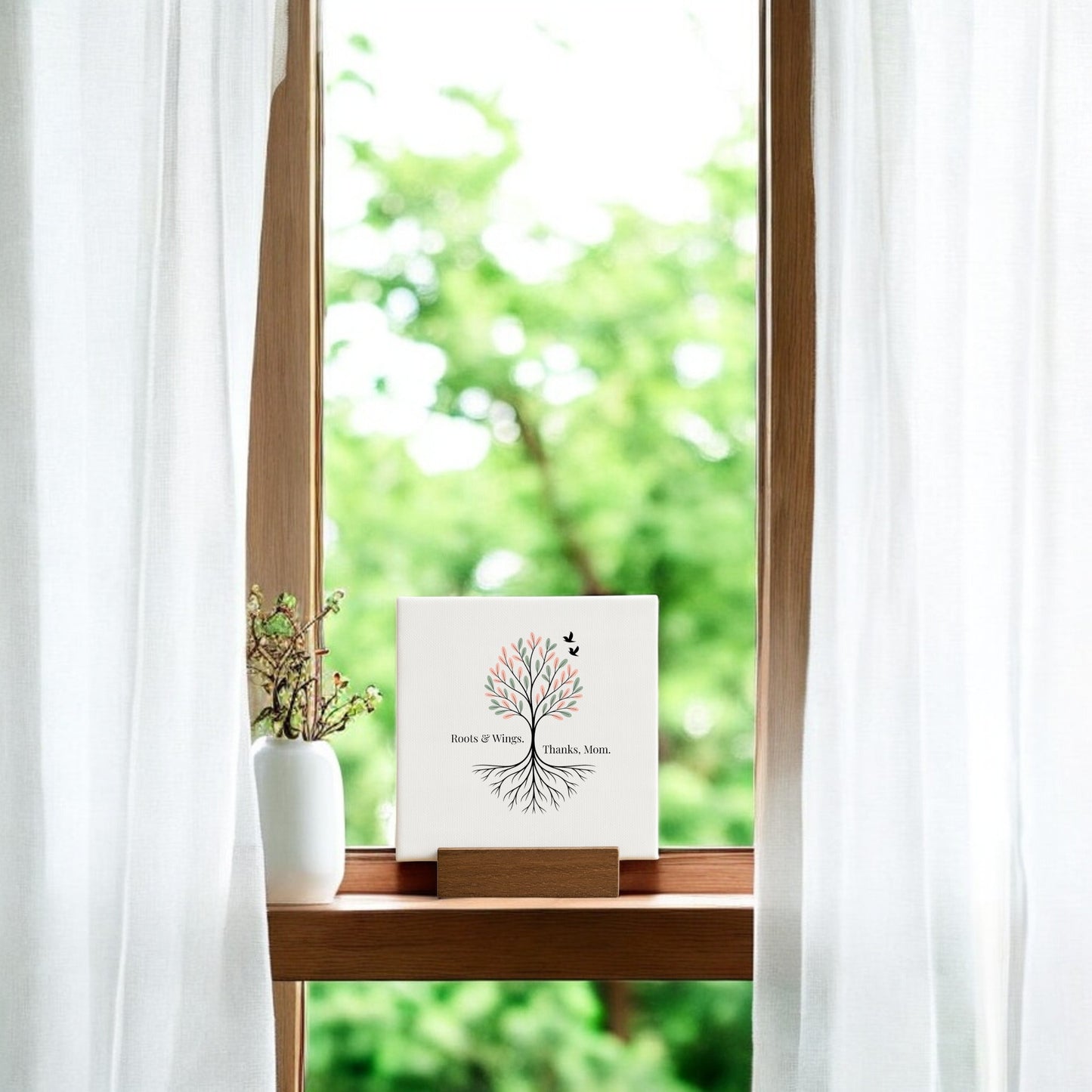 A thoughtful gift for Mom, the Roots & Wings Mini Sign – Mother's Day Tree Canvas by Mirabilia Boutique features a tree design and “Roots of Home. Thanks, Mom.” It stands on a windowsill beside a vase with branches and white curtains.
