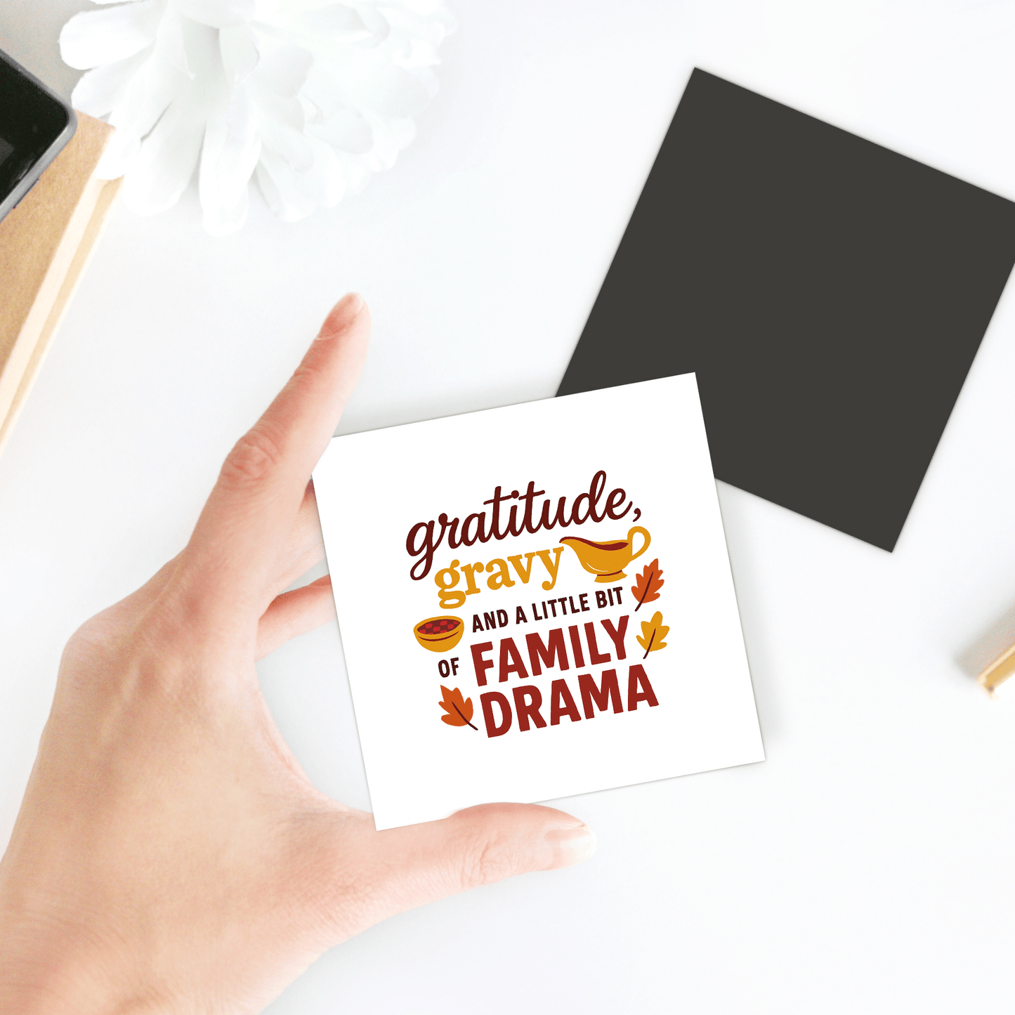 A hand holds Mirabilia Boutique's "Gratitude Gravy & Family Drama" magnet, a funny fall decor piece with leaf and gravy art. In the background, a white flower and black square rest on a white surface.