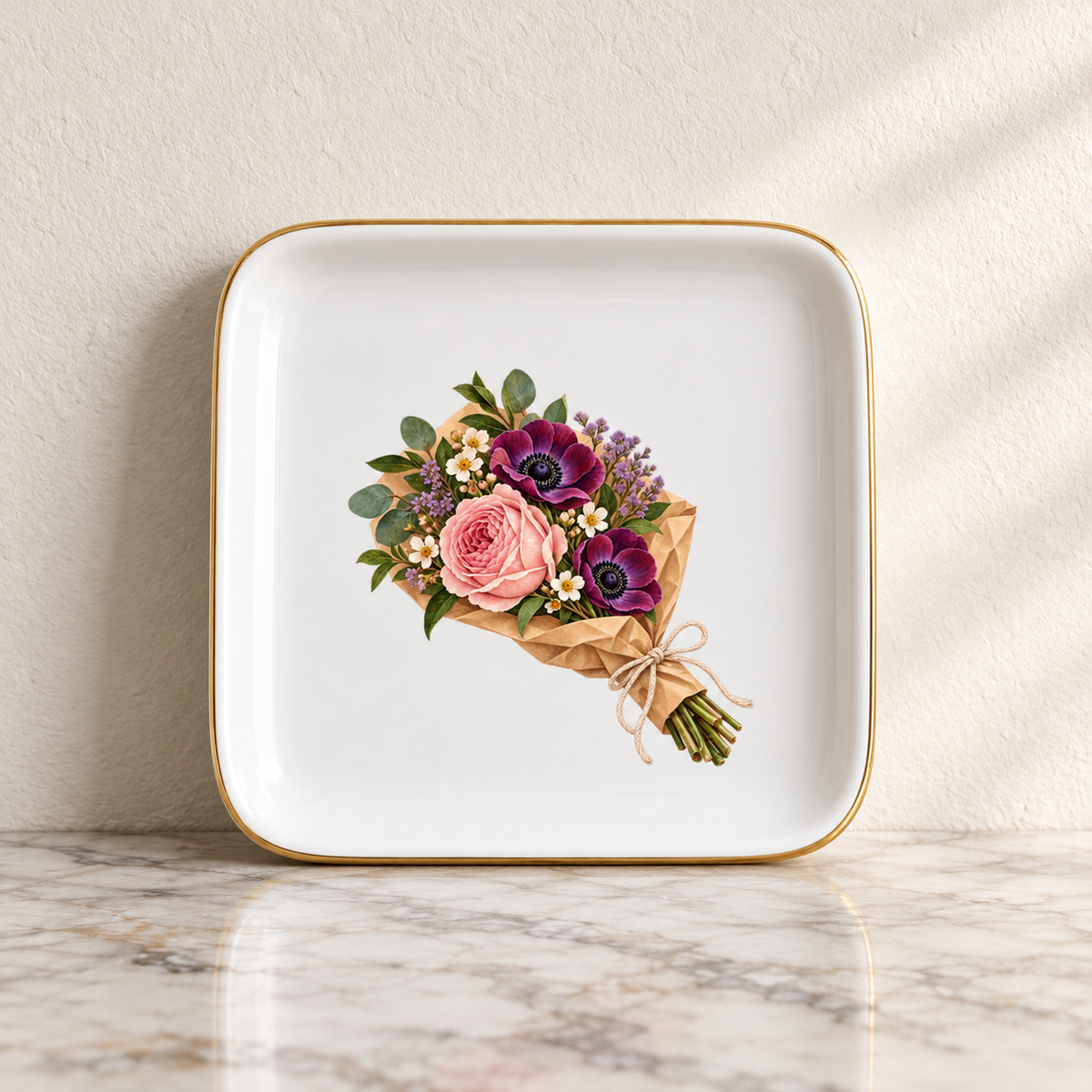 The Florist’s Parcel Jewelry Tray by Mirabilia Boutique is a square white ceramic tray with a gold rim, featuring an illustration of pink roses, purple anemones, and greenery wrapped in brown paper.