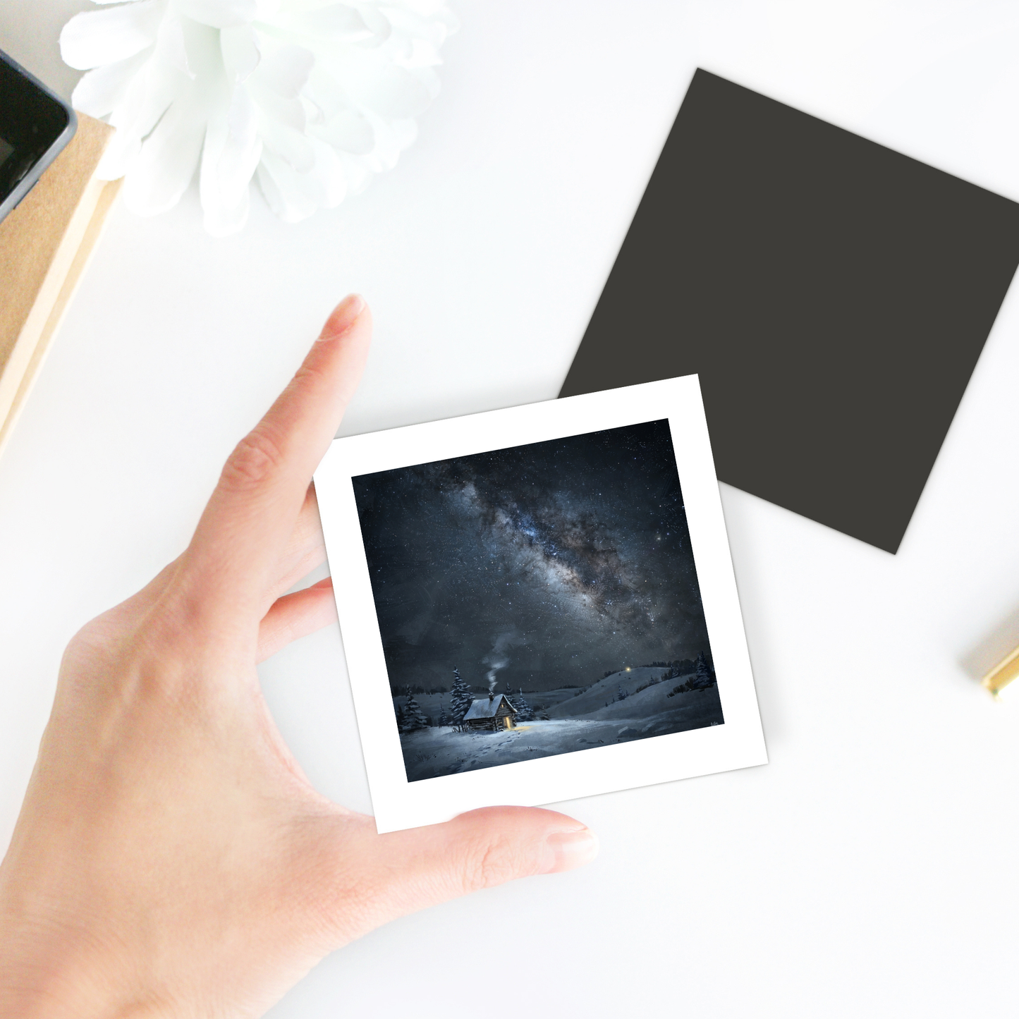 A hand holds “The Night Is Listening Milky Way Magnet | Celestial Art” by Mirabilia Boutique, featuring a square image of a snowy cabin beneath a starry night sky. On the white table in the background, there is also a flower, a black square, and a pencil.
