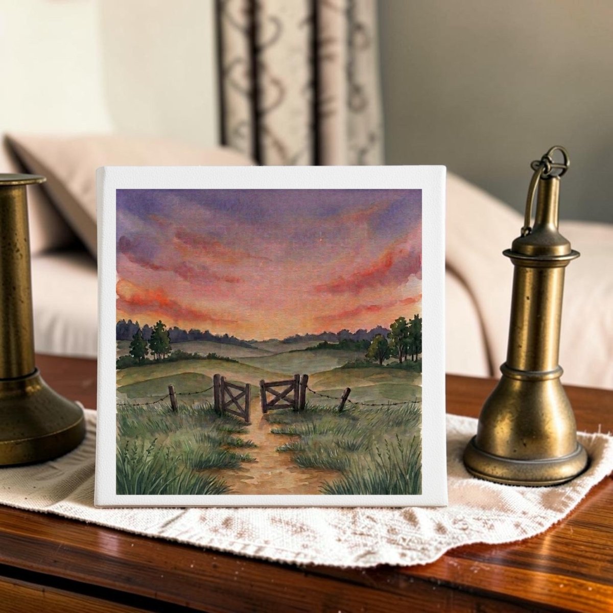 The “There Is More Sunset Landscape Canvas Art” (8x8) by Mirabilia Boutique, featuring a country path with a wooden gate and fence leading to grassy fields beneath a vibrant sunset sky, is displayed on a lace cloth between two brass candlesticks atop a wooden table.