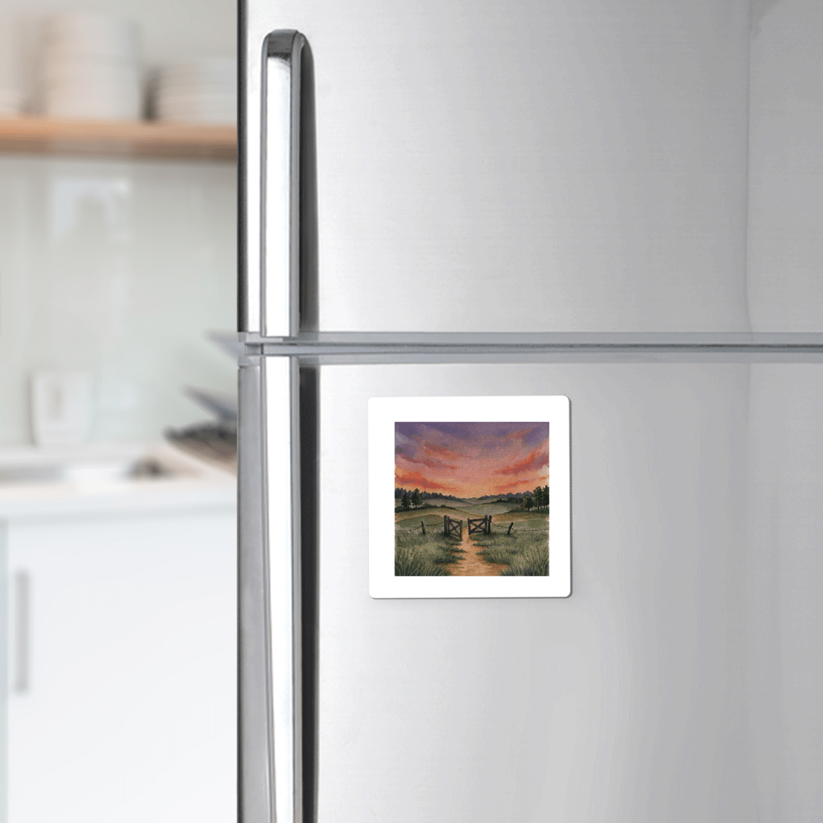 A “There Is More Sunset Landscape Magnet | Inspirational Art” by Mirabilia Boutique, featuring a rural sunset scene with a country path and wooden gate, is displayed on a stainless steel refrigerator in a modern kitchen.