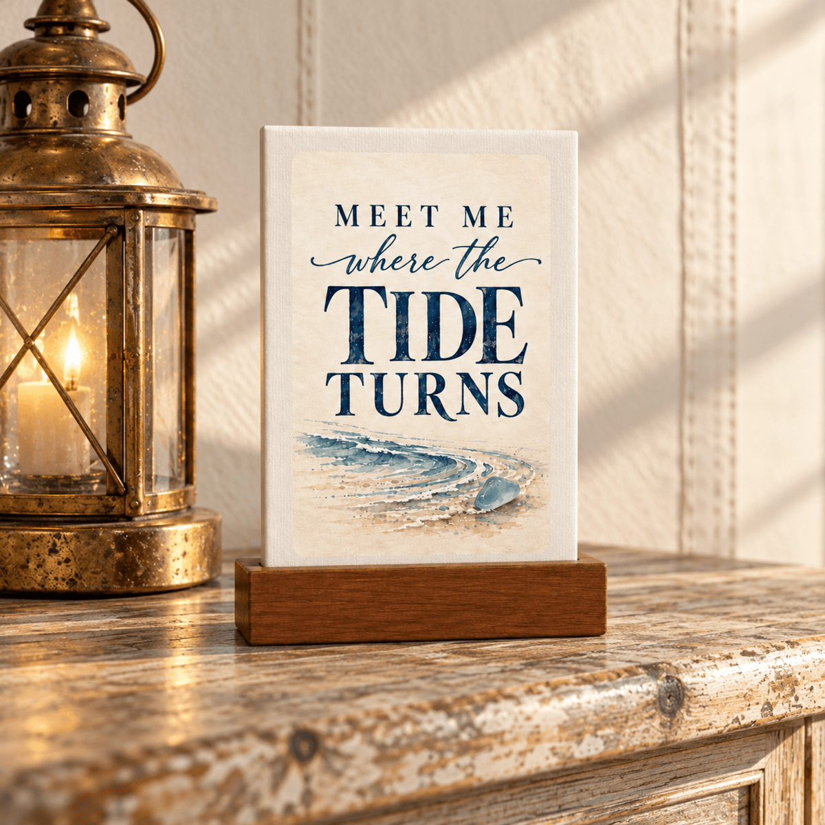 A Meet Me Where the Tide Turns Mini Sign | Salt & Story rests in a wooden stand above a watercolor shoreline painting, set next to a lit metal lantern as part of a sea glass decor display.