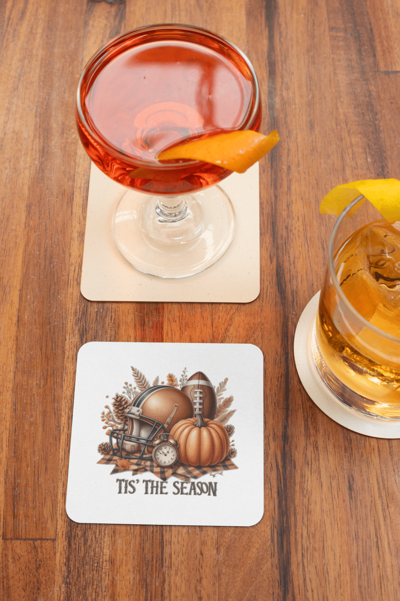 Two cocktails sit on the Tis’ the Season Coaster Set by Mirabilia Boutique. A third football-themed coaster features a helmet, pumpkin, autumn leaves, and “Tis’ the Season”—the perfect fall football drink decor for your next game day.