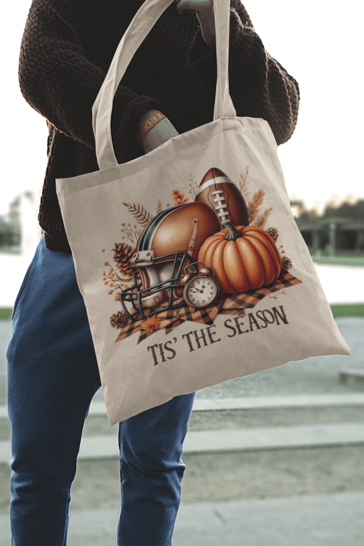 A person holds the "Tis’ the Season Tote Bag" by Mirabilia Boutique, a fall market bag with football, helmet, pumpkin, vintage clock, autumn leaves, and "TIS’ THE SEASON" printed underneath.